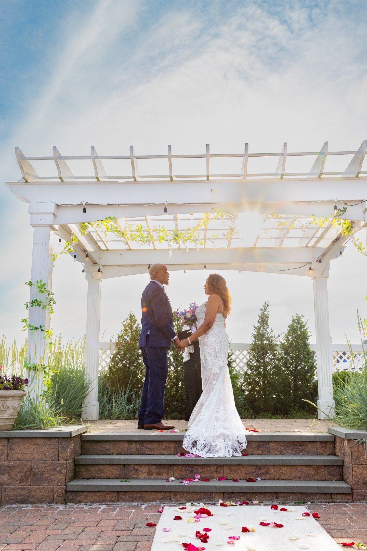 A bride and groom holding hands and gazing at each other during their wedding ceremony on an outdoor stage with steps, surrounded by greenery and decorated with flower petals and plants.