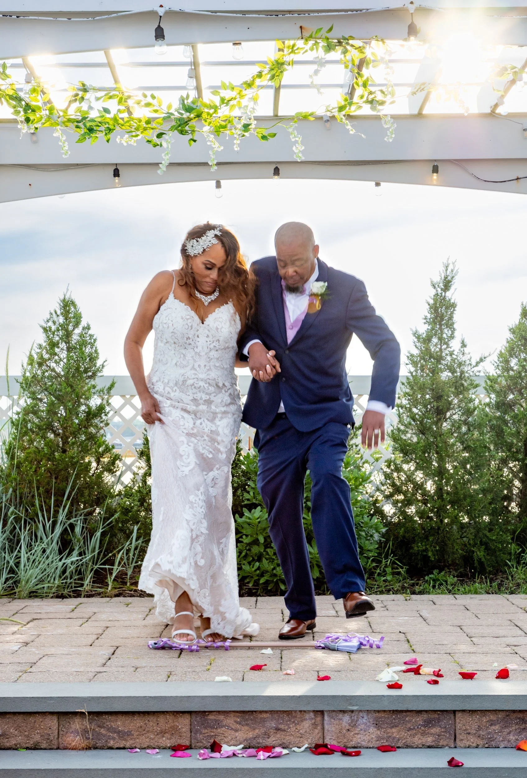 A couple jumps the broom together beneath a pergola, with rose petals scattered on the steps.