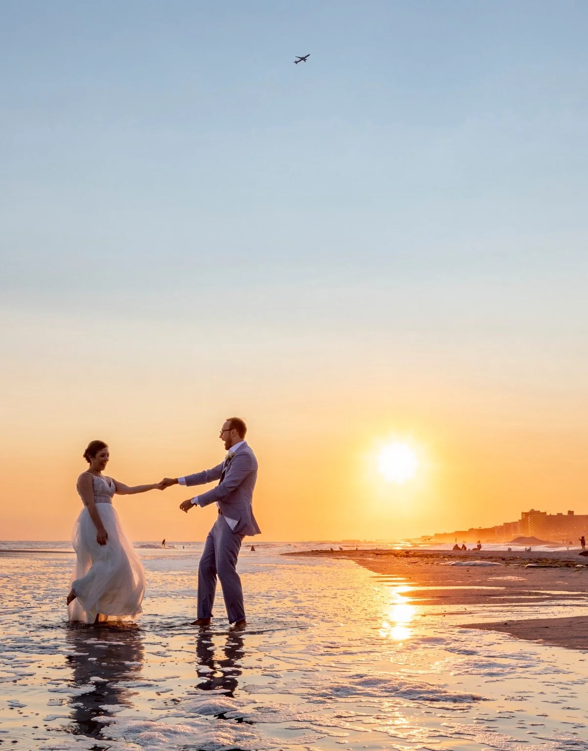 A couple holding hands in shallow ocean water at sunset, with the sun and their reflections visible