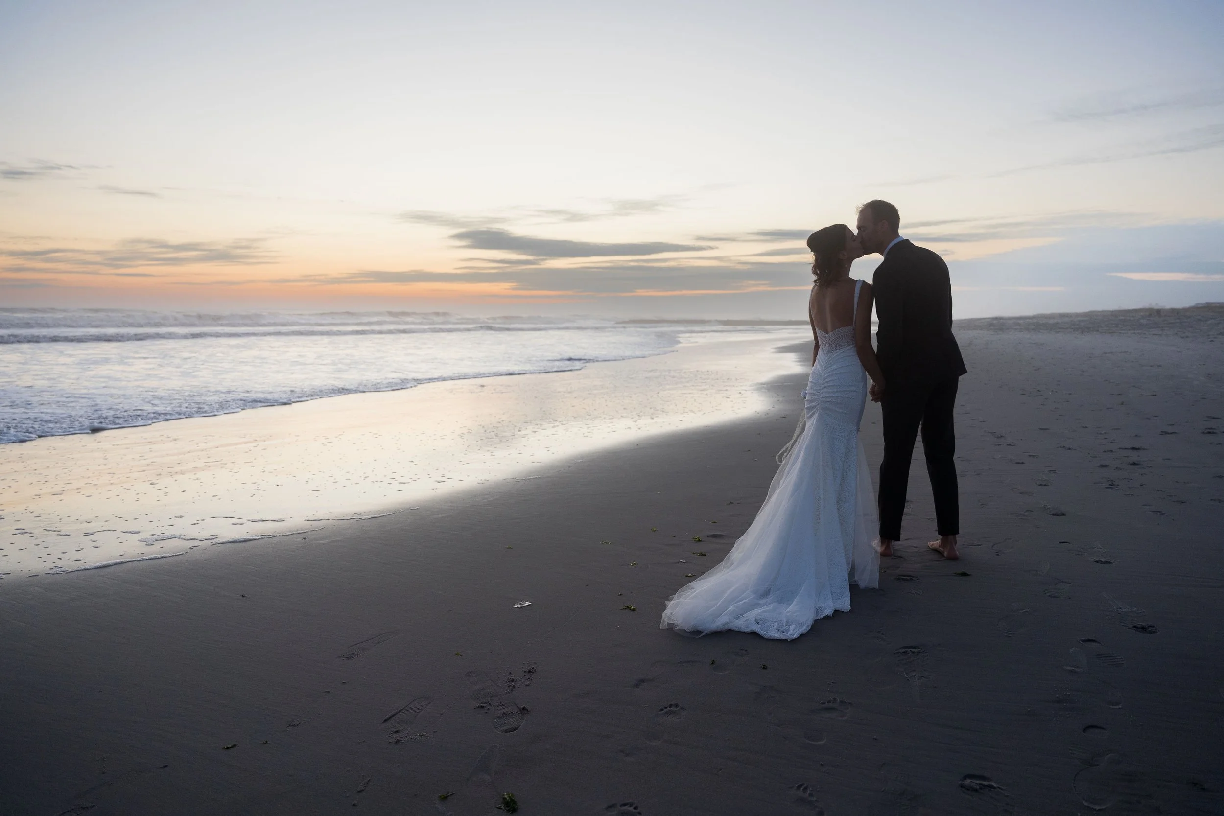 A bride and groom sharing a kiss on a beach at sunset, with the ocean in the background.