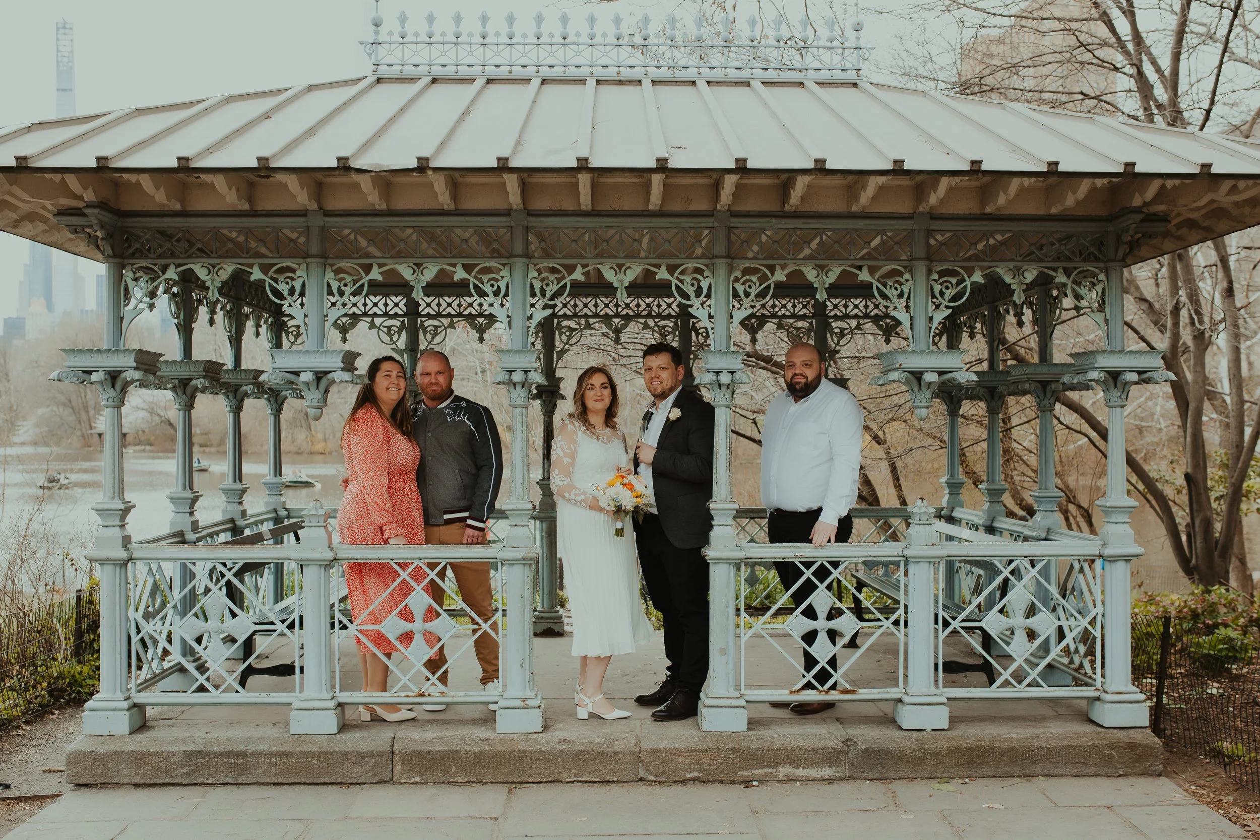 Group of five people standing inside a decorative gazebo in a park, celebrating a wedding, with the bride and groom in the center holding a bouquet and a glass, surrounded by trees and city buildings in the background.