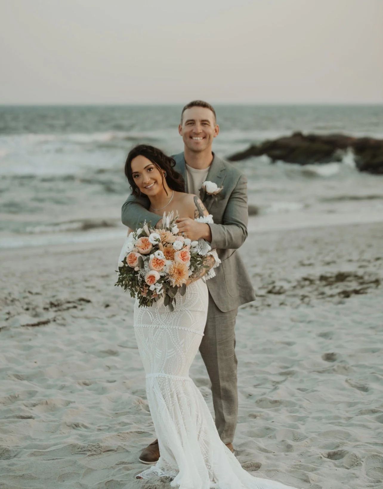A newlywed couple smiling on the beach, with the woman holding a bouquet of flowers and the man hugging her from behind.