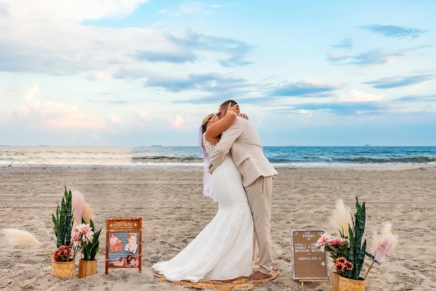 A couple in wedding attire embraces on a beach during sunset or late afternoon. The woman wears a white lace wedding dress, and the man wears a beige suit. There are decorative plants and signs on the sand, one sign reads "The wedding of Kayla & Daur August 9, 2022." The scene is romantic with a calm ocean and sky with clouds in the background.