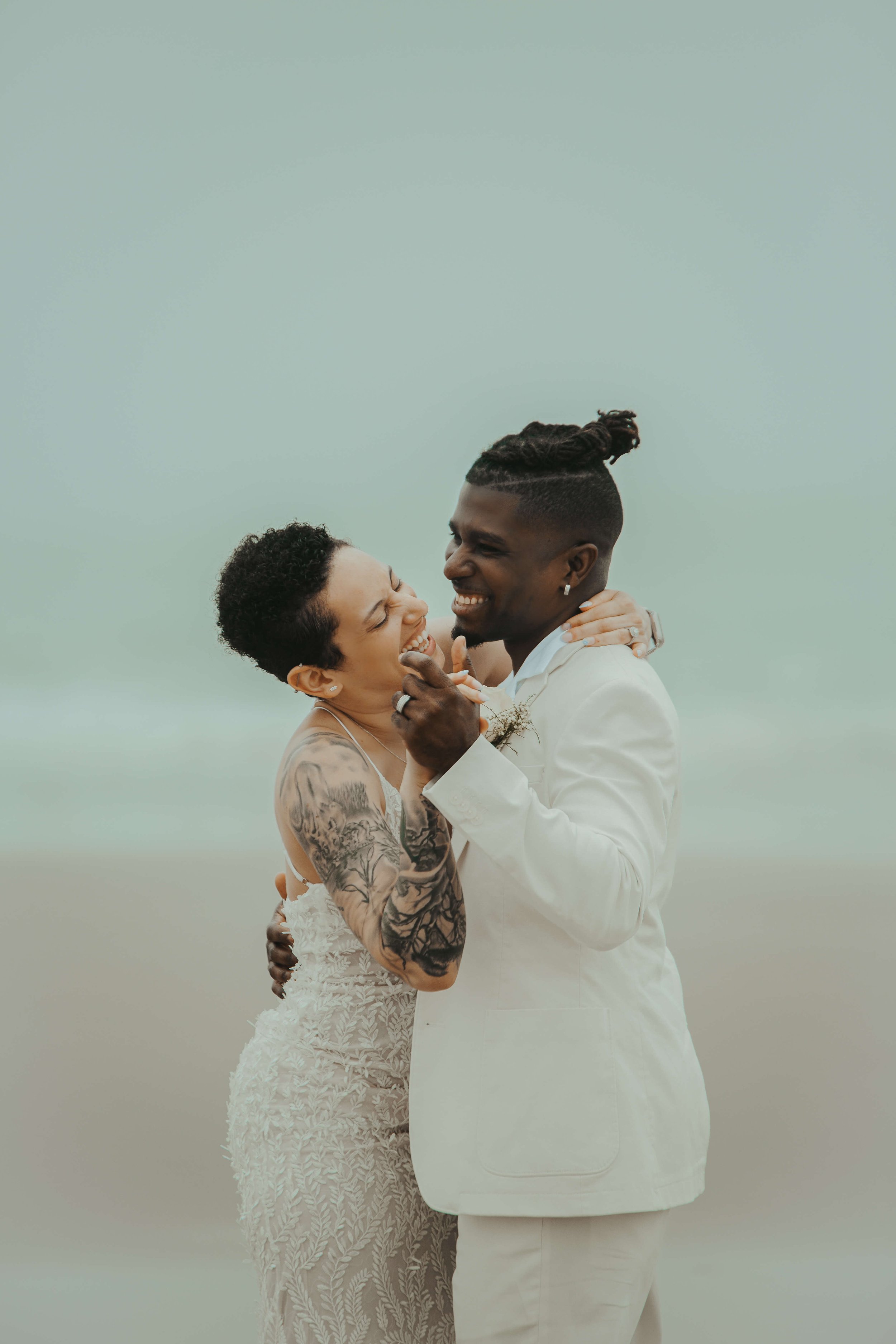A happy couple is embracing and laughing on a beach, dressed in wedding attire.