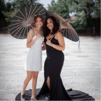 Two women standing outdoors under black umbrellas, smiling at the camera. One is wearing a white dress, and the other is in a black gown.
