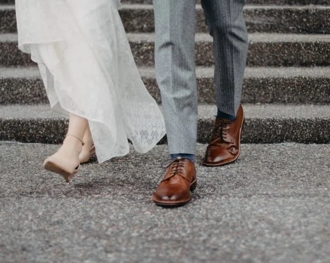 Close-up of a bride and groom walking down concrete stairs, showing their lower bodies and shoes. The bride is wearing a white wedding dress and beige heels, and the groom is dressed in gray pants and brown leather shoes.