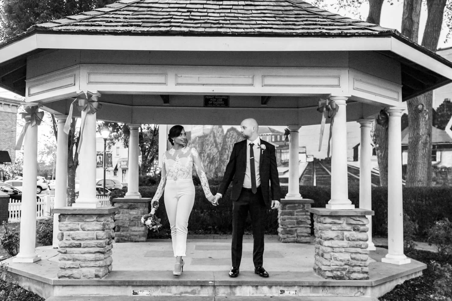 A couple, dressed in wedding attire, holding hands under a small pavilion, surrounded by trees and parked cars.