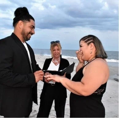 A man and woman exchange rings during a beach wedding ceremony, with a officiant standing behind them, under a cloudy sky.