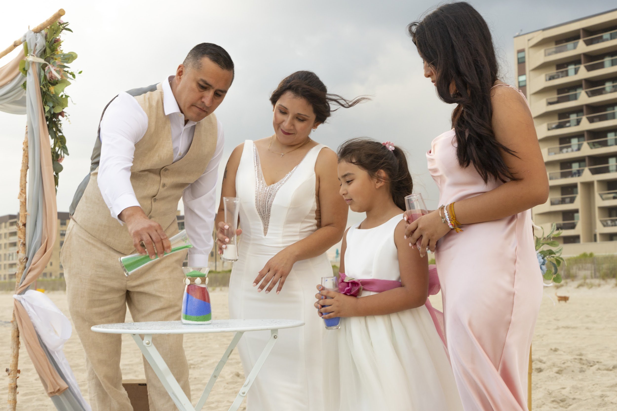 A couple and their family pour colored sand into one vase during a beach sand ceremony.