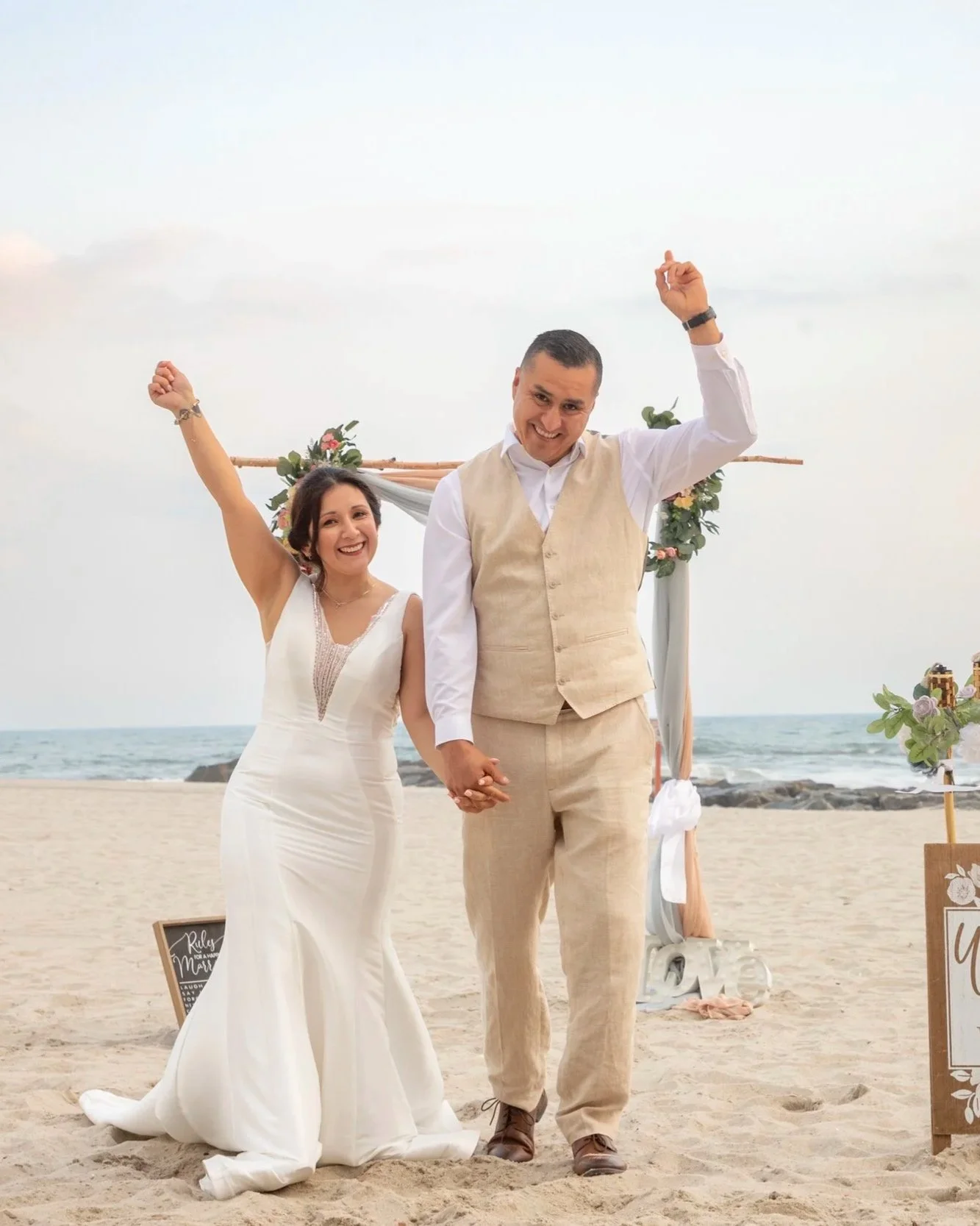 A smiling couple in wedding attire holding hands on a beach, with a decorated wooden arch and signs in the background.