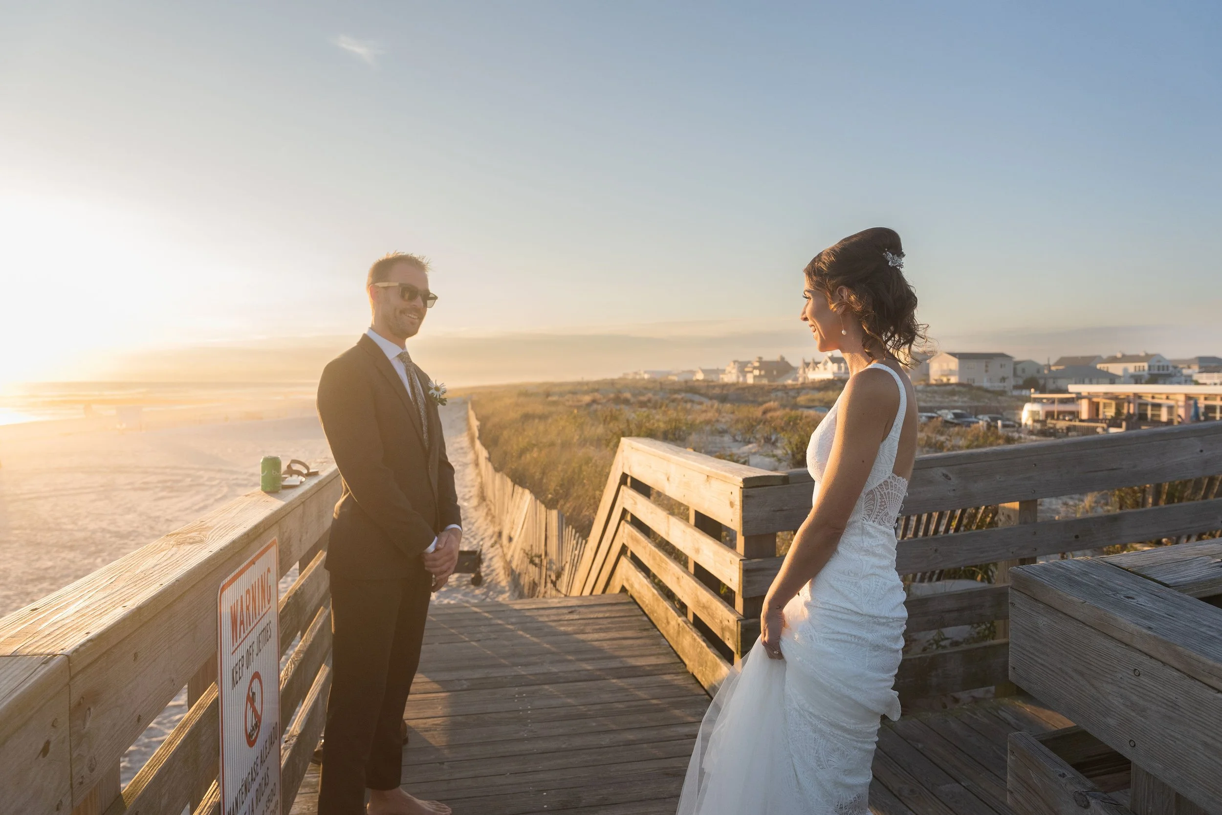 A bride and groom standing on a wooden beach pier at sunset, smiling and facing each other, with houses and dunes in the background.