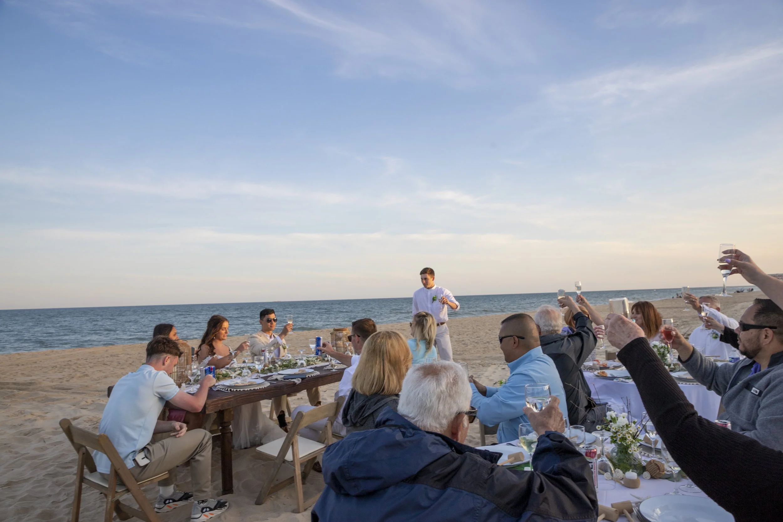 People gather on a beach for a celebration, raising glasses in a toast, with the ocean and sky in the background during sunset.
