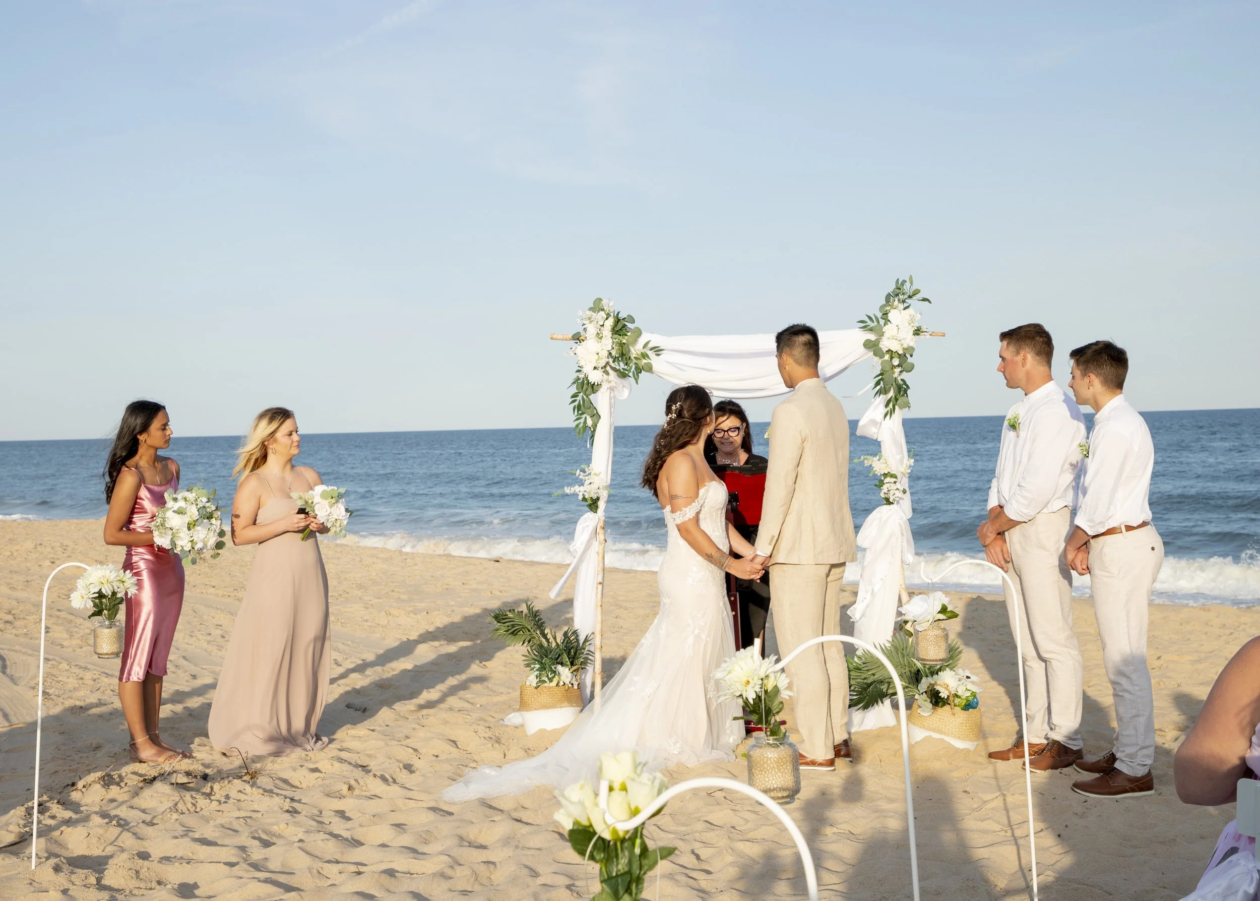 A wedding ceremony on the beach with a couple standing under a decorated arch, surrounded by bridesmaids and groomsmen, with the ocean and blue sky in the background.
