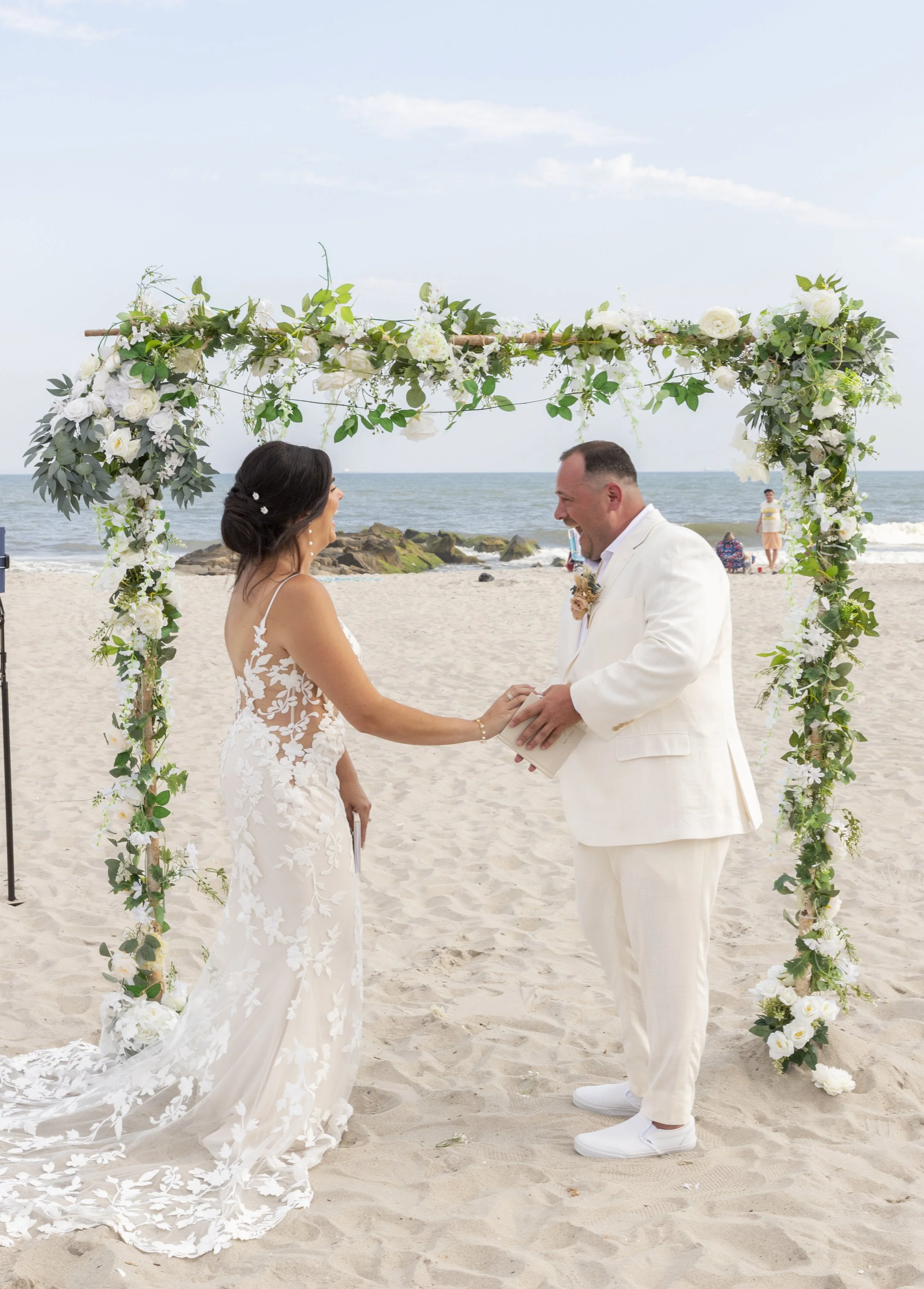 A bride and groom exchanging vows during a beach wedding ceremony under a floral arch, with the ocean in the background.