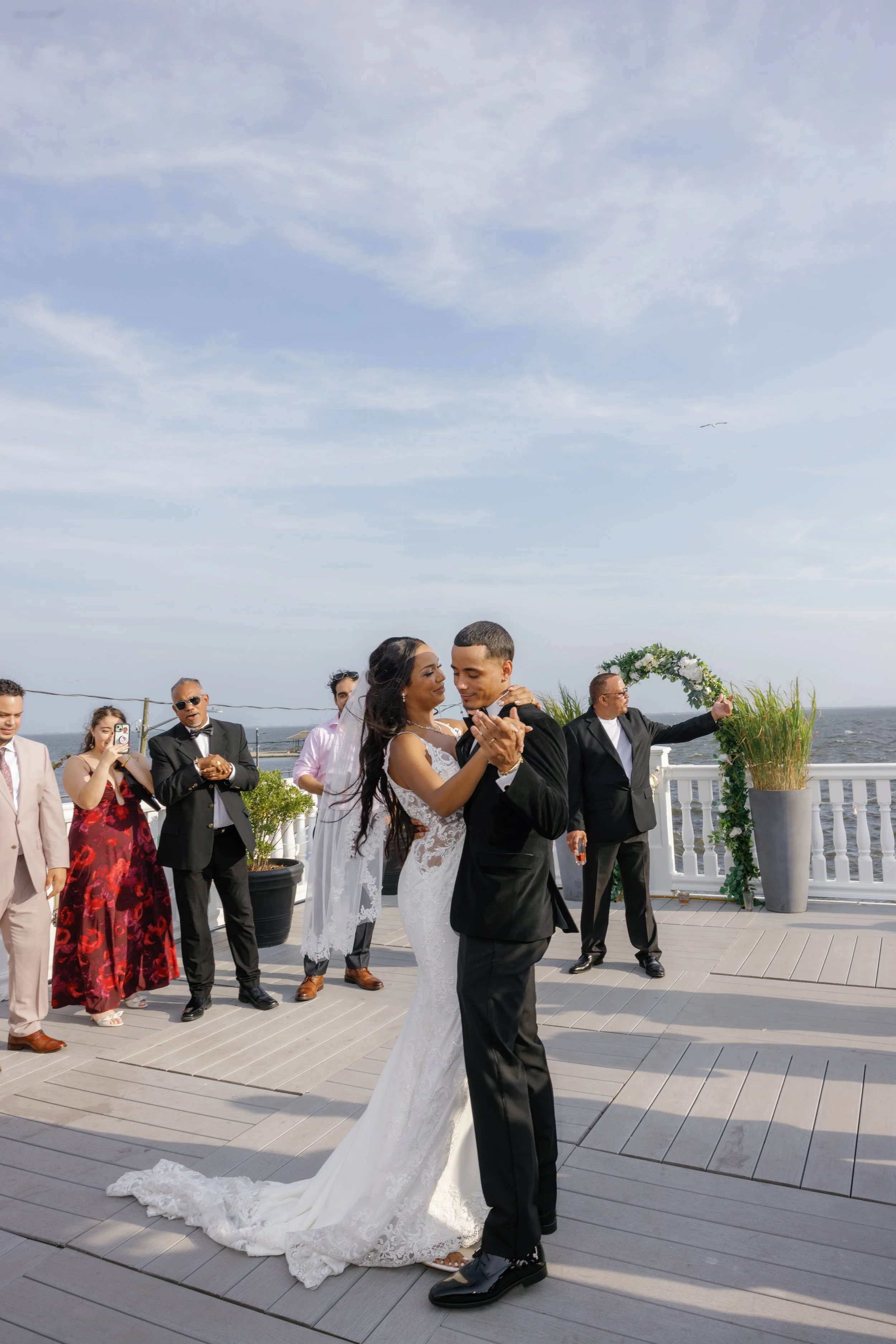 A bride and groom dance during their wedding on an outdoor deck, with wedding guests watching and a scenic ocean view in the background.