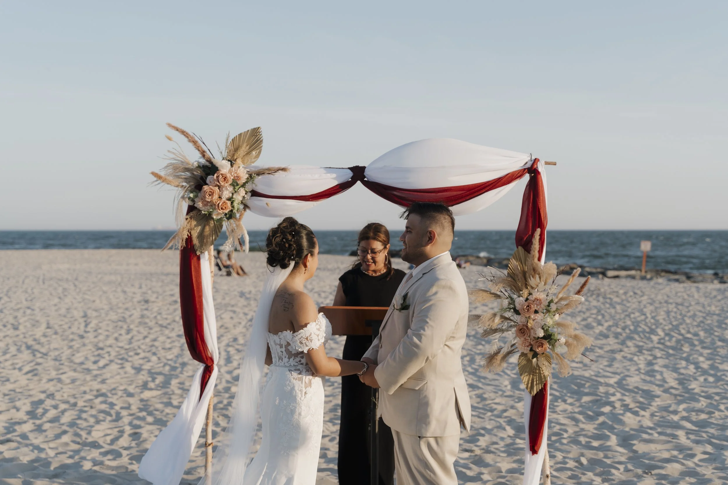 A wedding ceremony taking place on a beach with a bride and groom holding hands and facing each other under a decorated arch with flowers and fabric, with the ocean in the background.