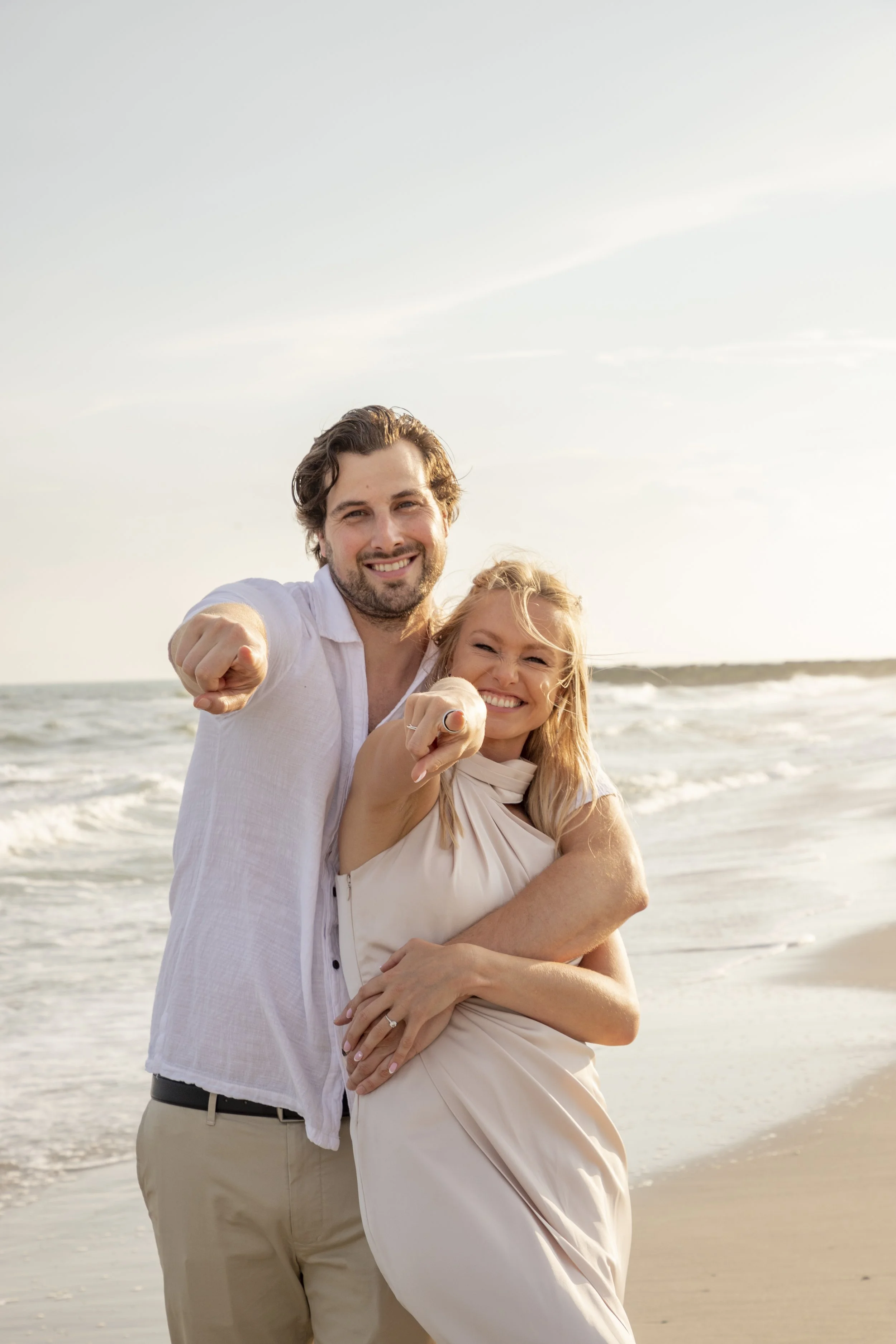 A smiling couple standing on the beach near the ocean, pointing at the camera, with waves in the background.