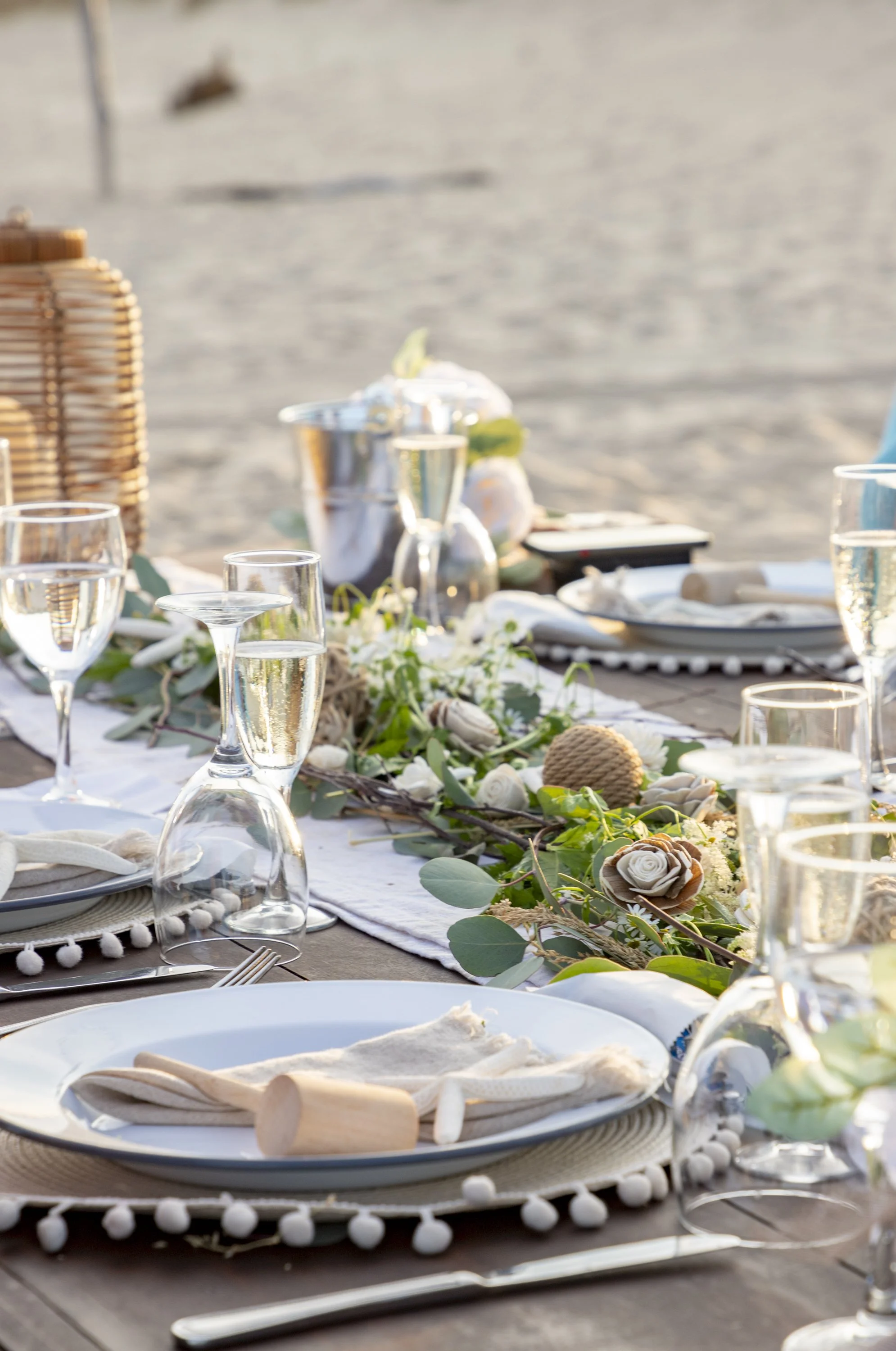 Elegant beachside dining table with glassware, plates, napkins, and floral centerpiece on sandy beach.