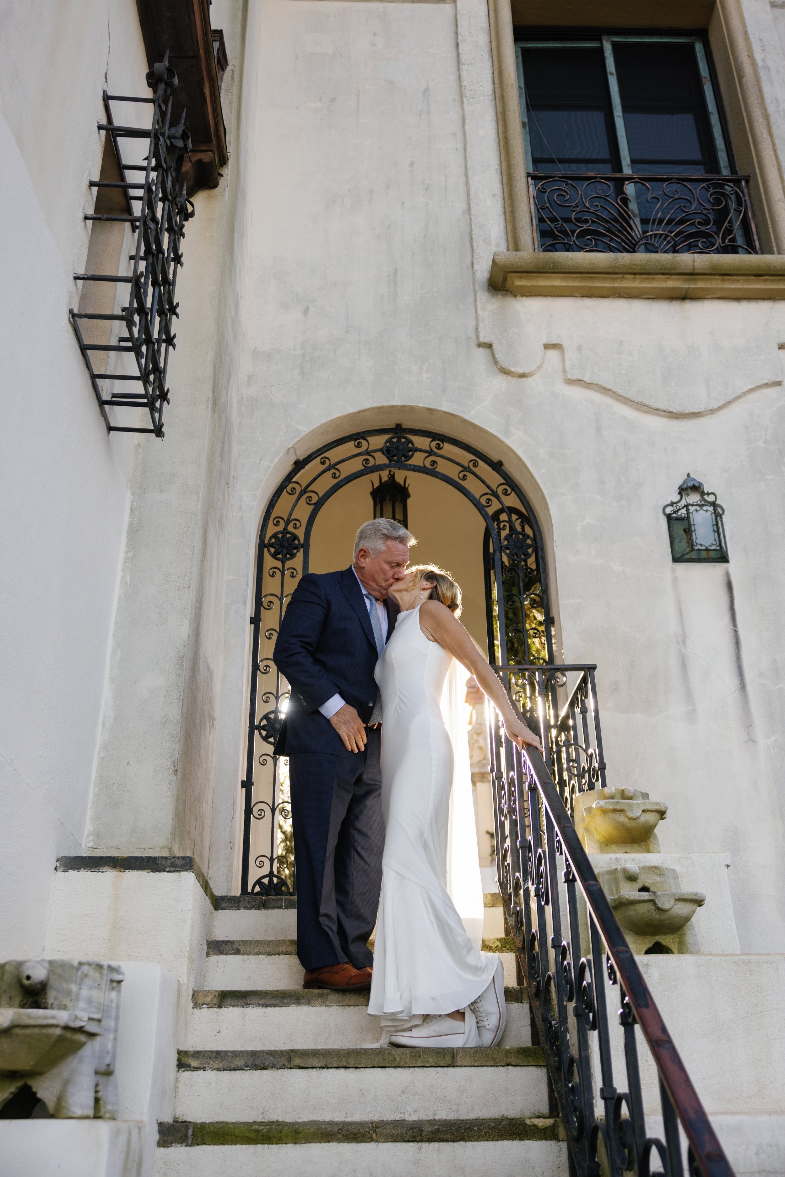 A bride and an older groom sharing a kiss on a staircase outside a building with ornate ironwork on the railing and a large arched doorway.