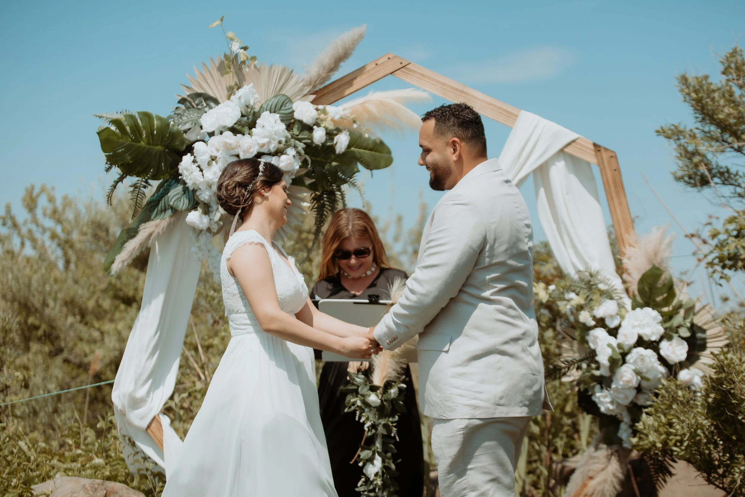 A couple getting married outdoors under a decorated wooden arch in Delray Beach, Florida with white flowers and green foliage, holding hands and smiling at each other, with an officiant smiling in the background and a clear blue sky overhead.