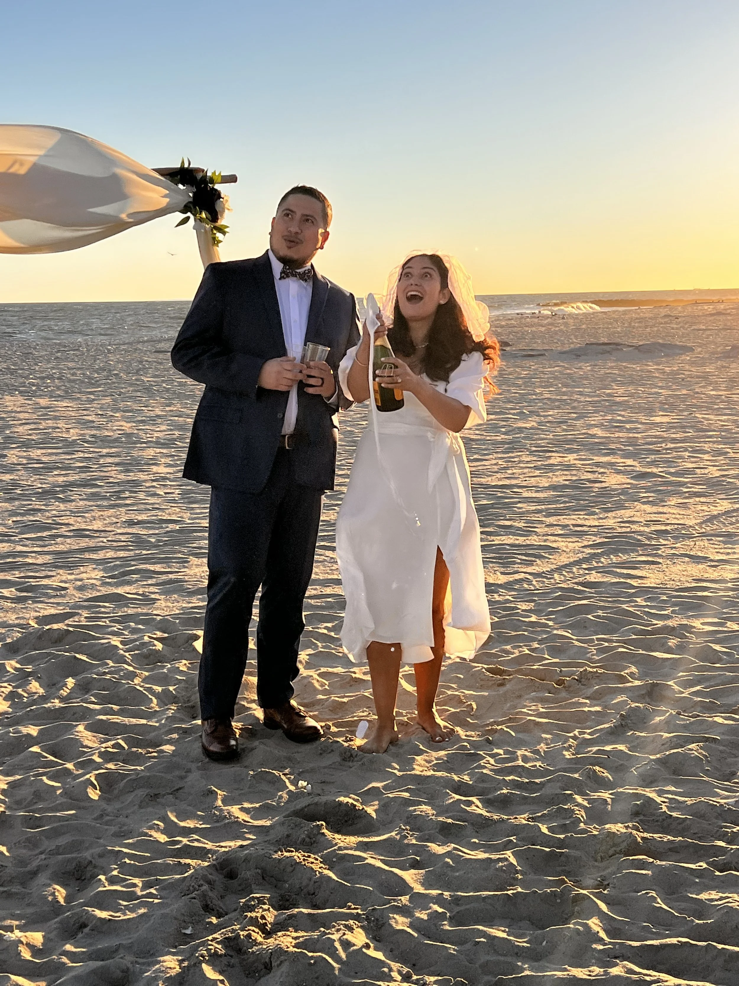 A couple dressed in wedding attire standing on the beach during sunset, celebrating with drinks in hand.