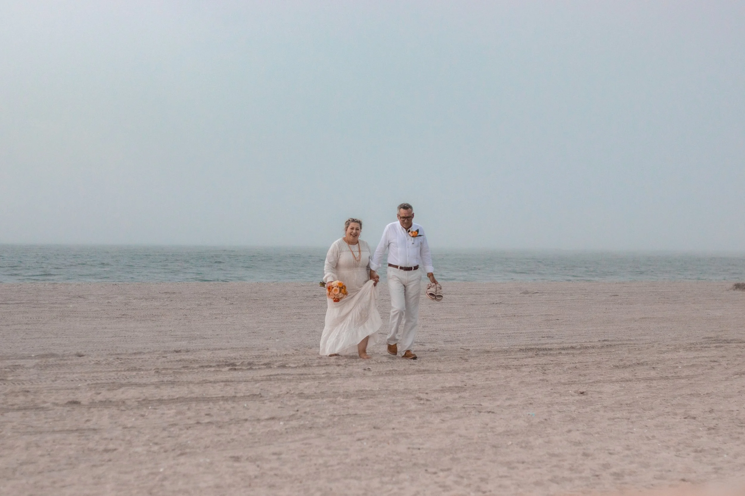 A couple in wedding attire walking barefoot on the beach, holding hands and smiling, with the ocean and sky in the background.