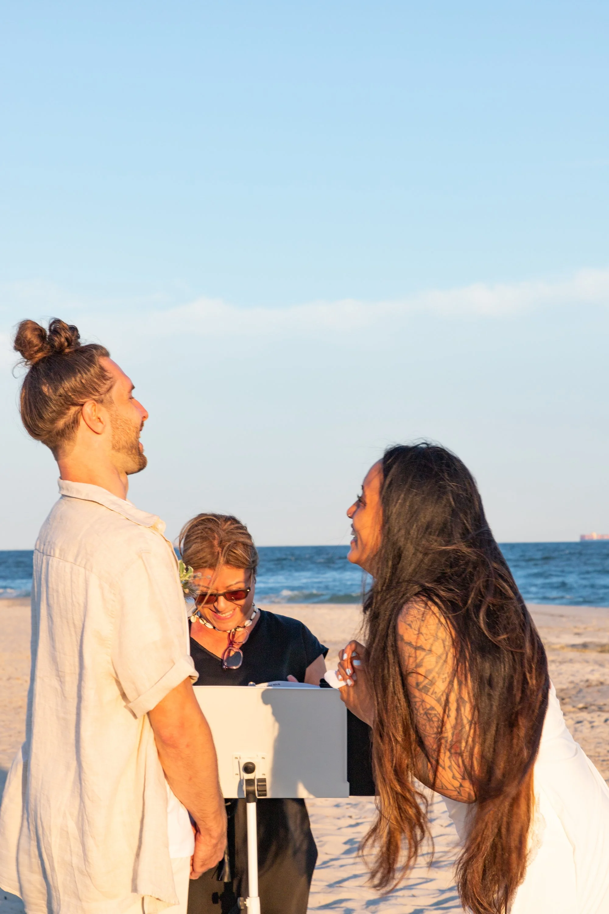 A couple getting married on the beach, exchanging vows, with a person officiating and holding a book.