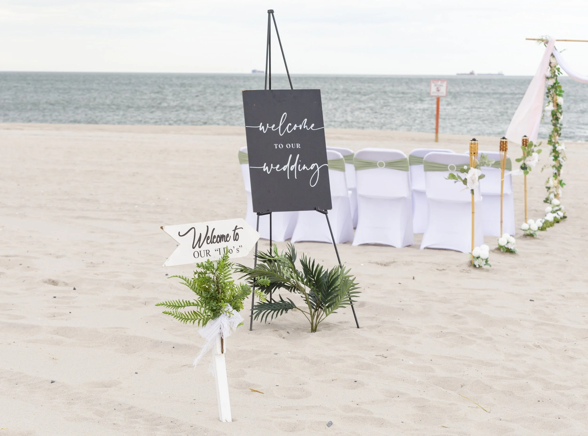 Beach wedding setup with decorated chairs facing the ocean, a black welcome sign on a stand, a white directional sign with greenery, and a floral arch in the background.