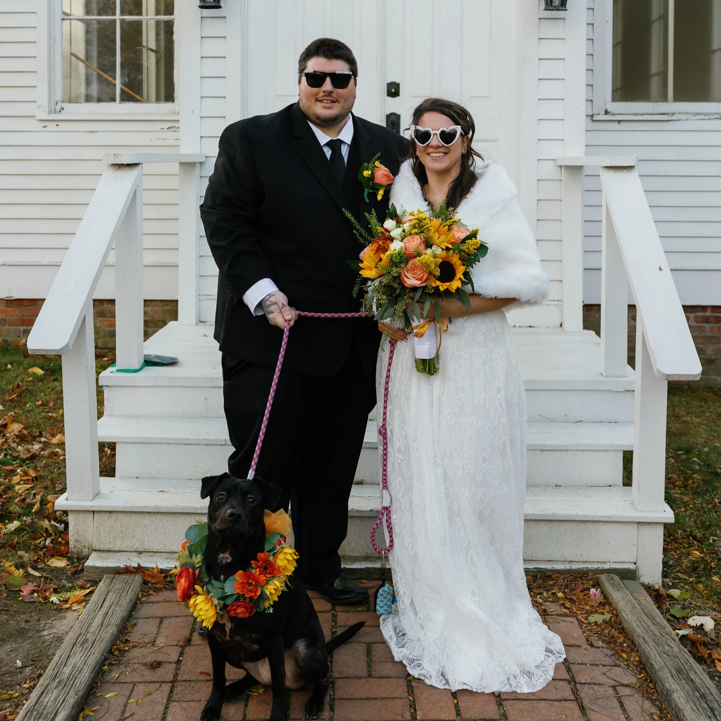 A couple dressed in wedding attire standing on the front steps of a house, holding a bouquet of flowers and a leash with a black dog wearing a flower wreath. The man is in a black suit and sunglasses, and the woman is in a white lace dress with heart-shaped sunglasses and a white shawl.