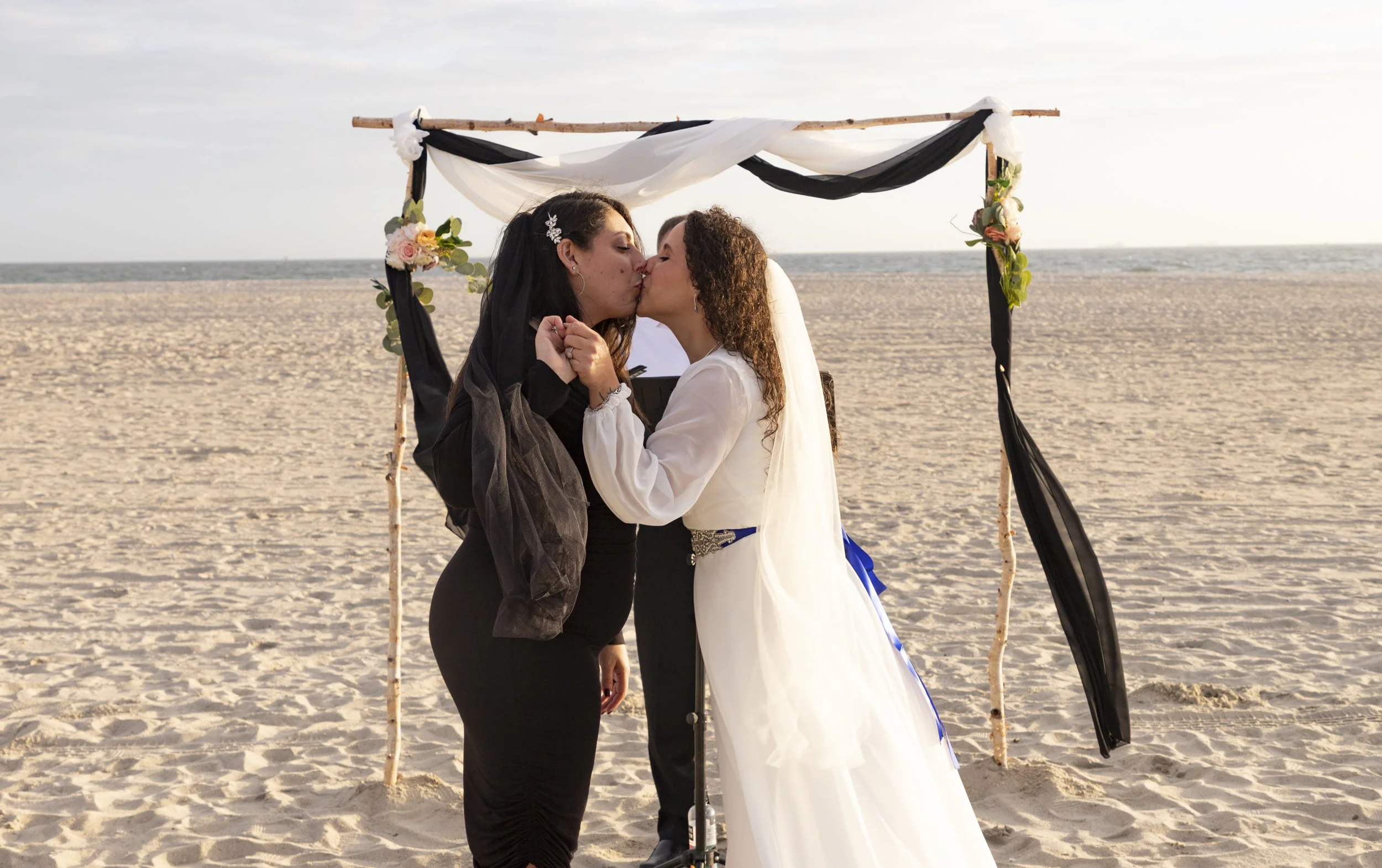 A couple sharing a kiss beneath a fabric-draped wedding arch on the beach.