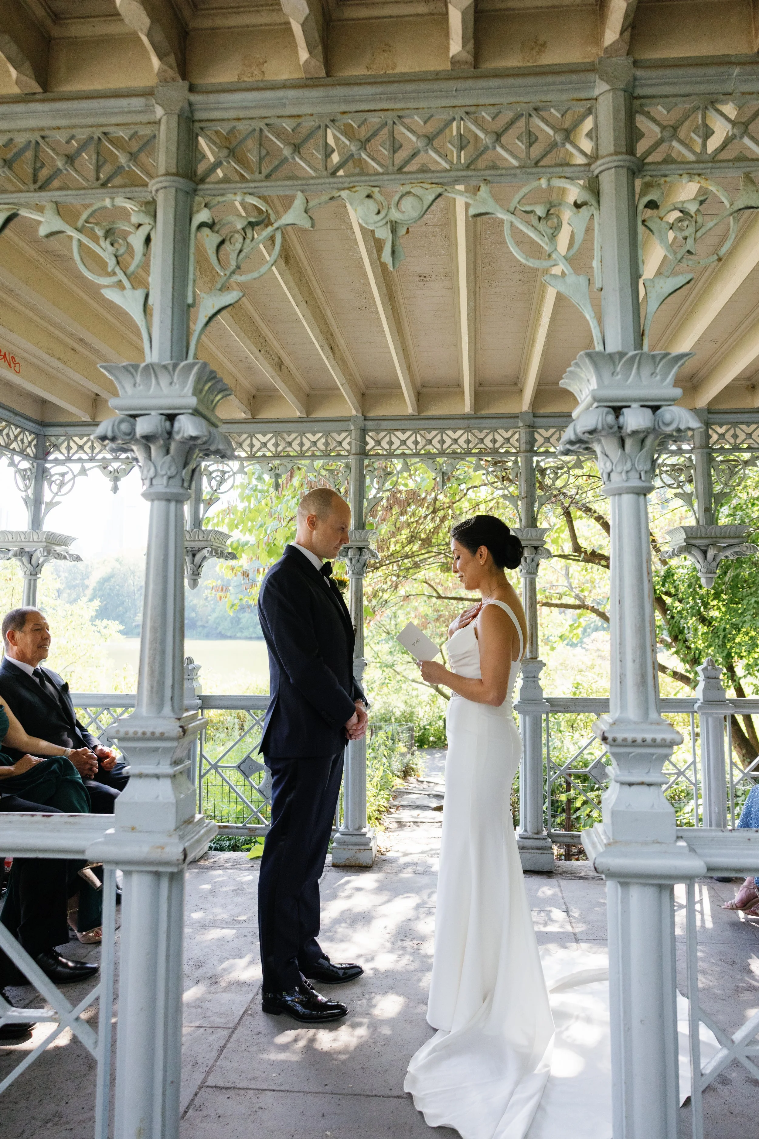 A couple exchanging vows during a wedding ceremony in a ornate gazebo outdoors, with two witnesses and lush greenery in the background.
