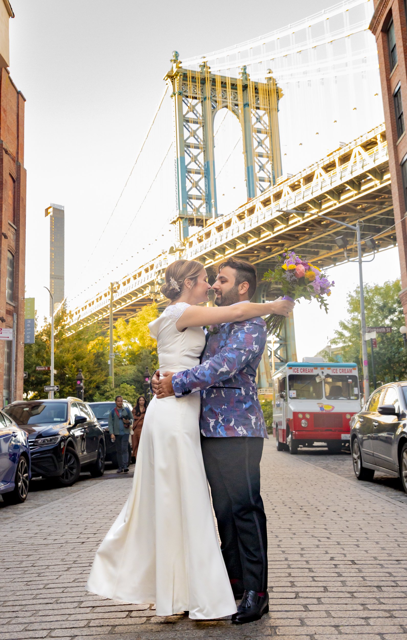 A couple dressed in wedding attire embracing on a city street with the Manhattan Bridge in the background, and a vintage ice cream truck nearby.