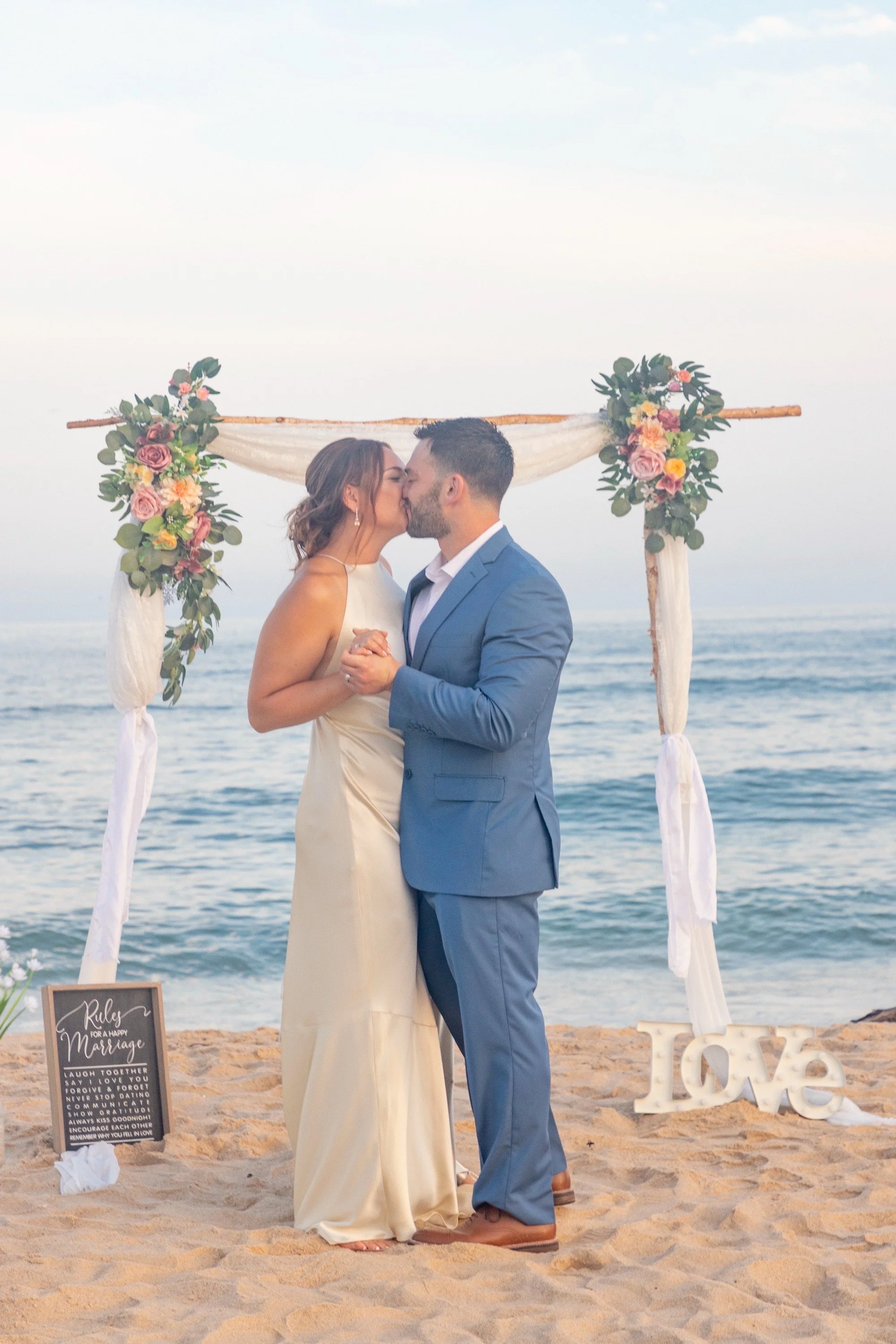 A couple kisses during their beach wedding ceremony at sunset, with an arch decorated with flowers and a sign reading 'Love' on the sand.