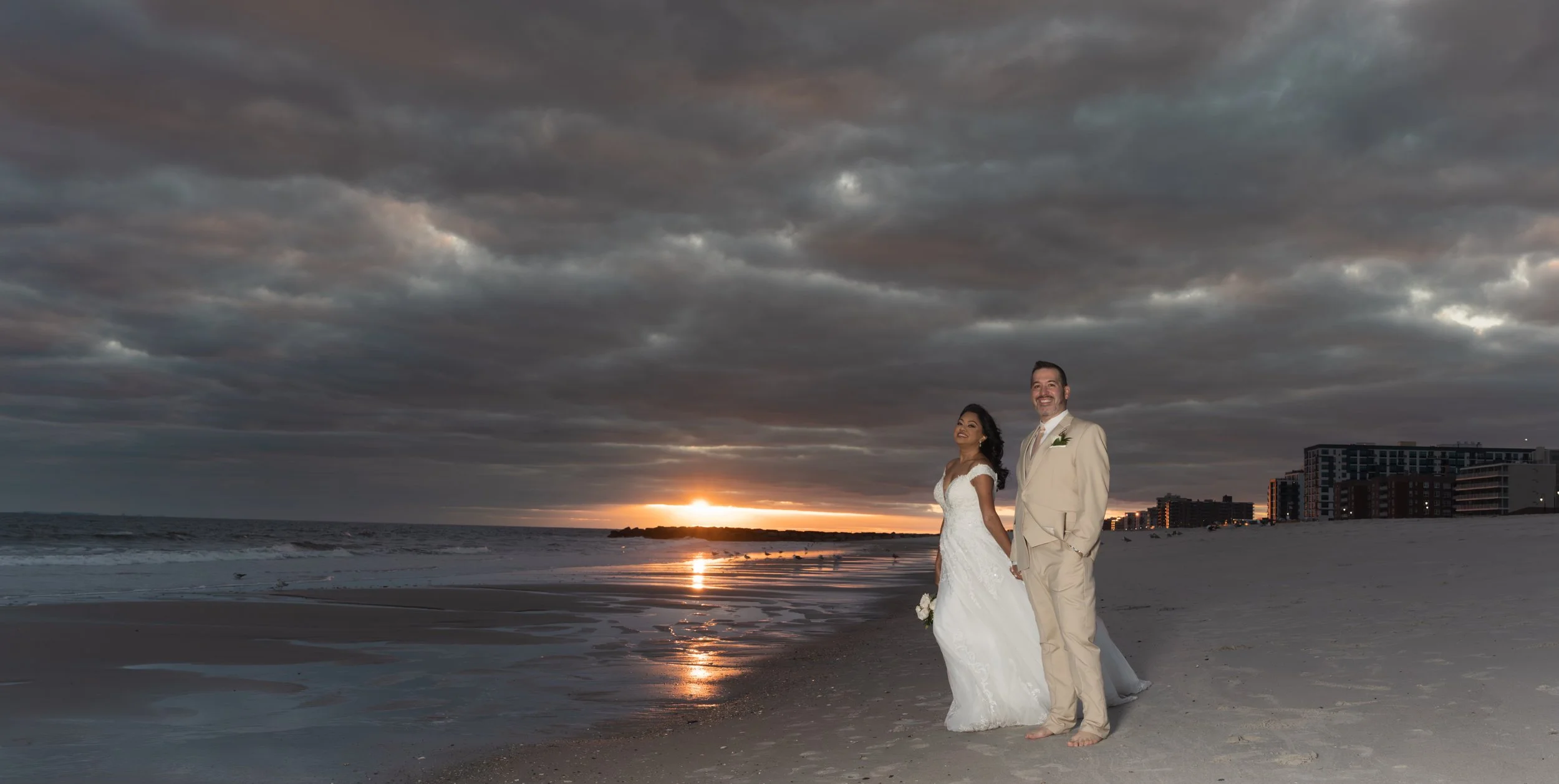 A bride and groom embrace on a rocky beach, touching foreheads, with the ocean and gray sky behind them.