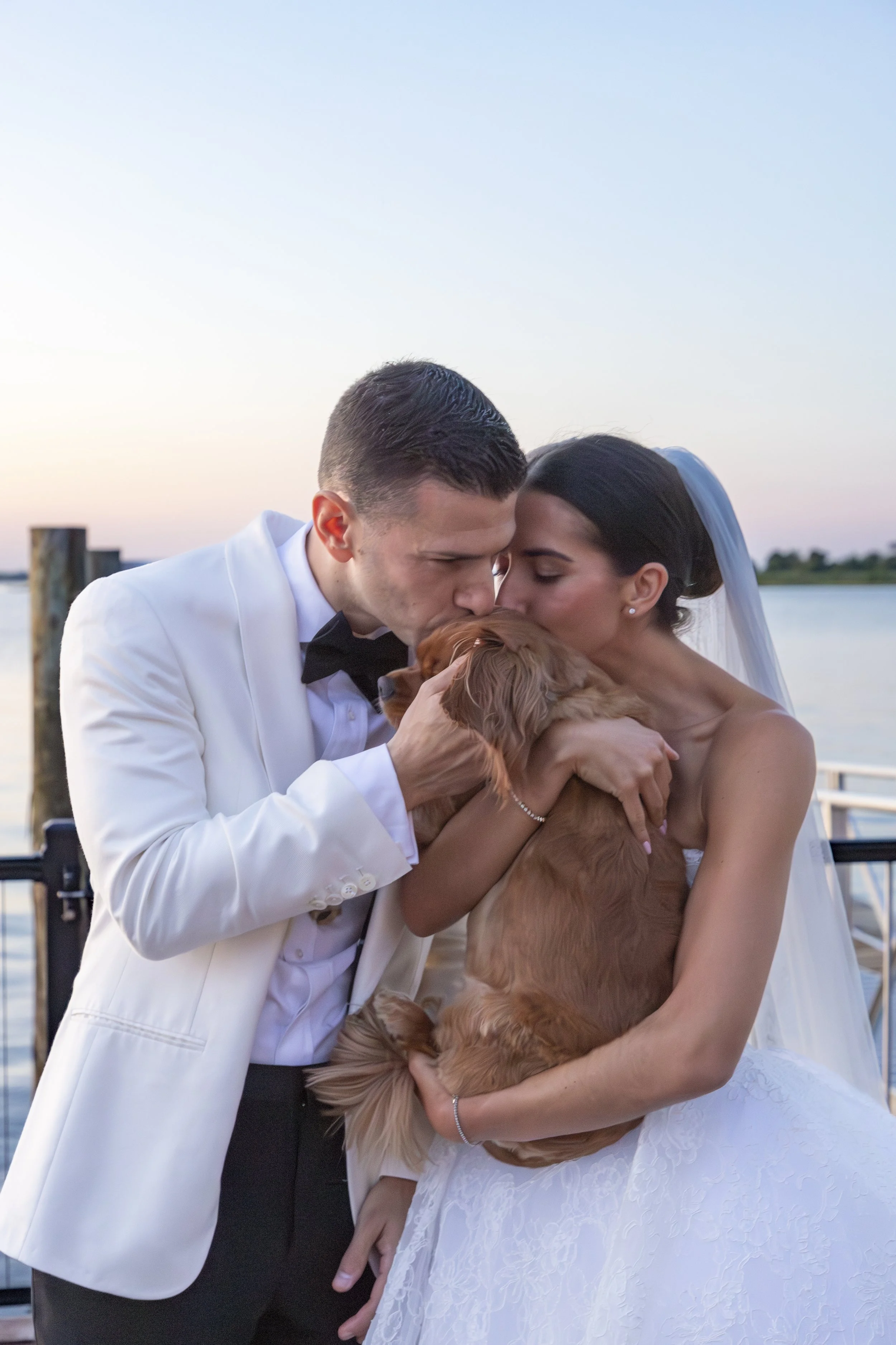 A newlywed couple kissing and holding a dog by the water during sunset.