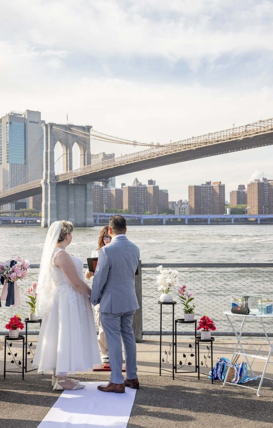 A wedding ceremony taking place outdoors near the water with the Brooklyn Bridge in the background, featuring a bride, groom, and officiant.