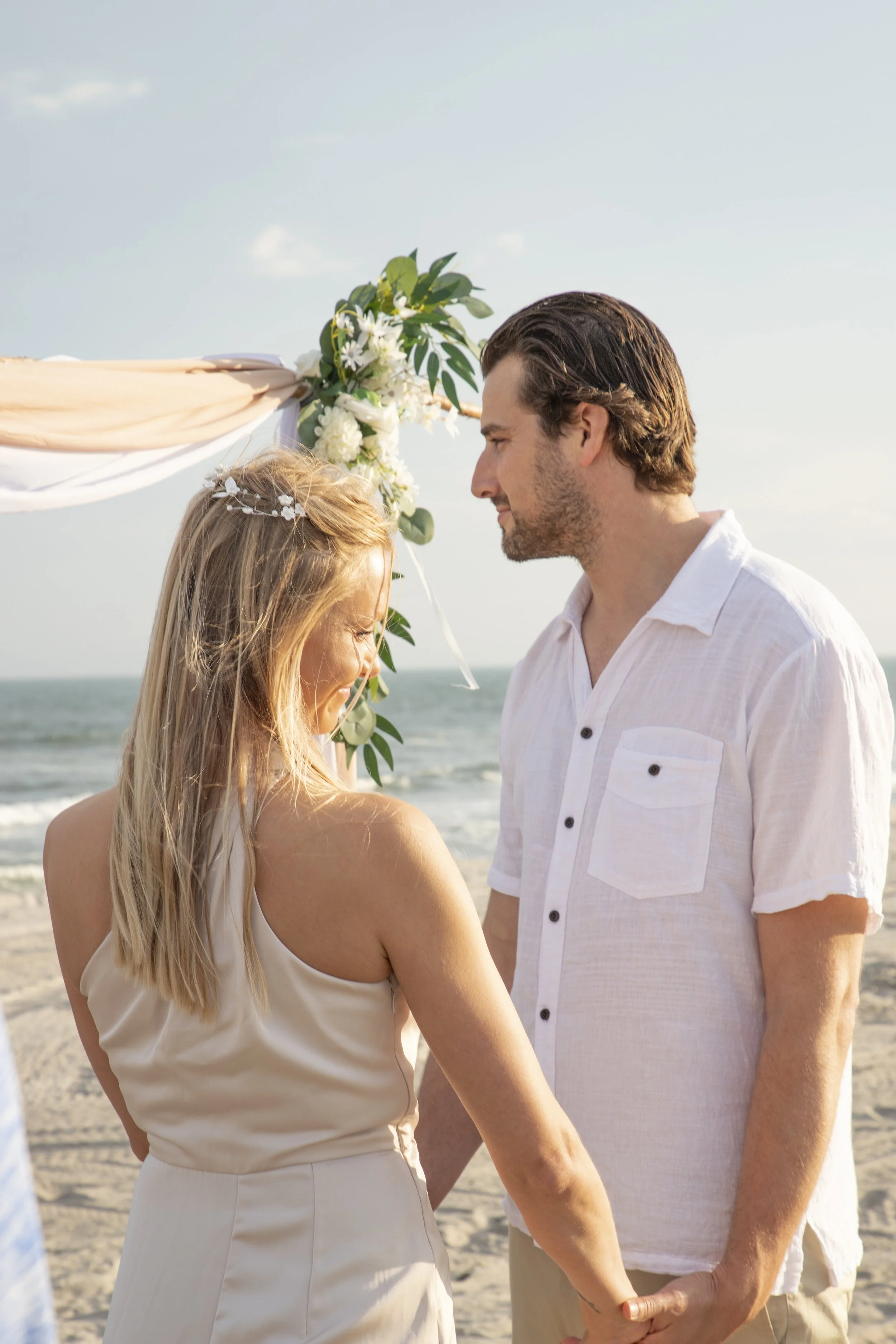 A couple holding hands, standing on the beach during their wedding ceremony, with an arch decorated with flowers and fabric behind them.