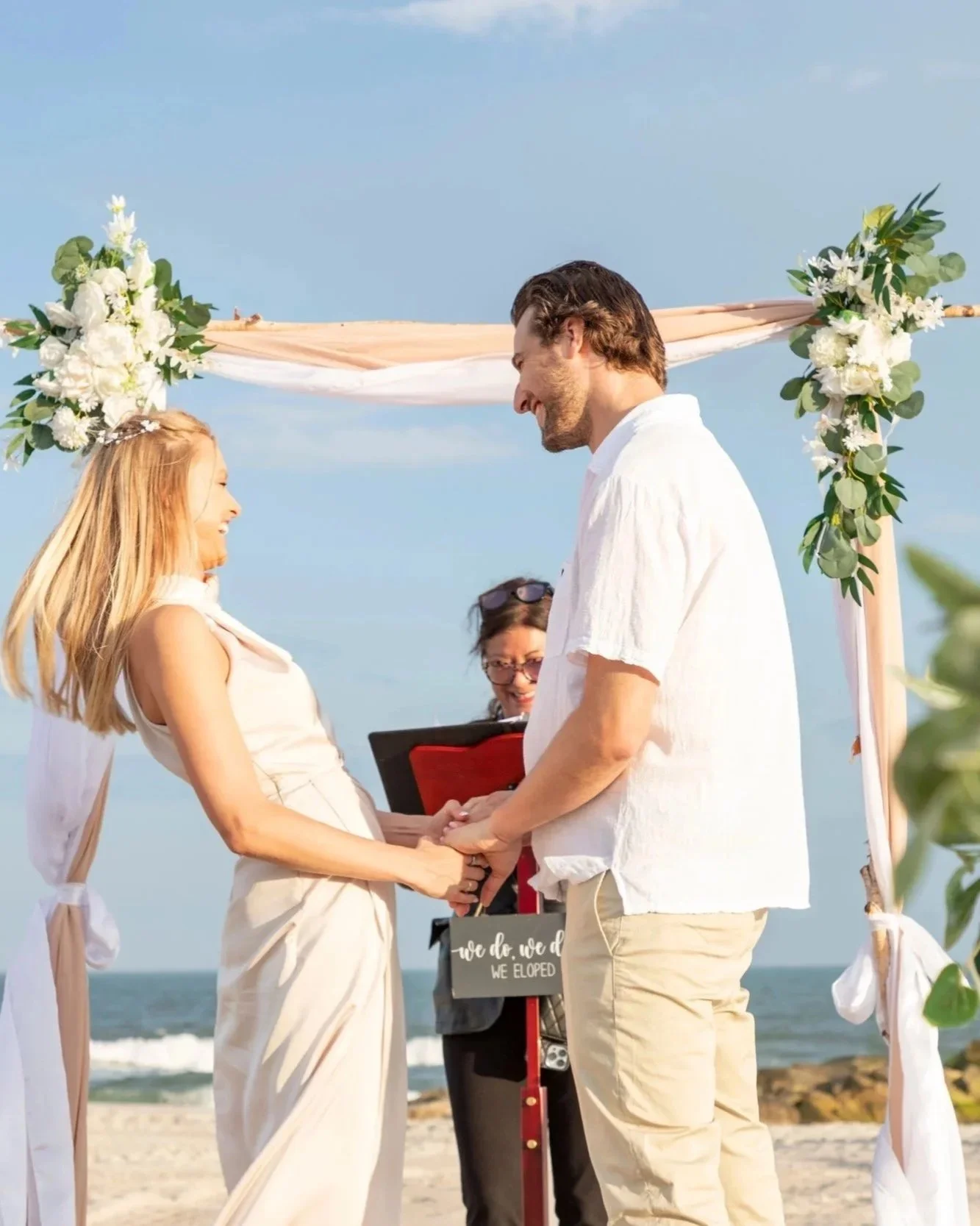 A couple getting married on the beach, holding hands and smiling at each other under a decorated beach arch.
