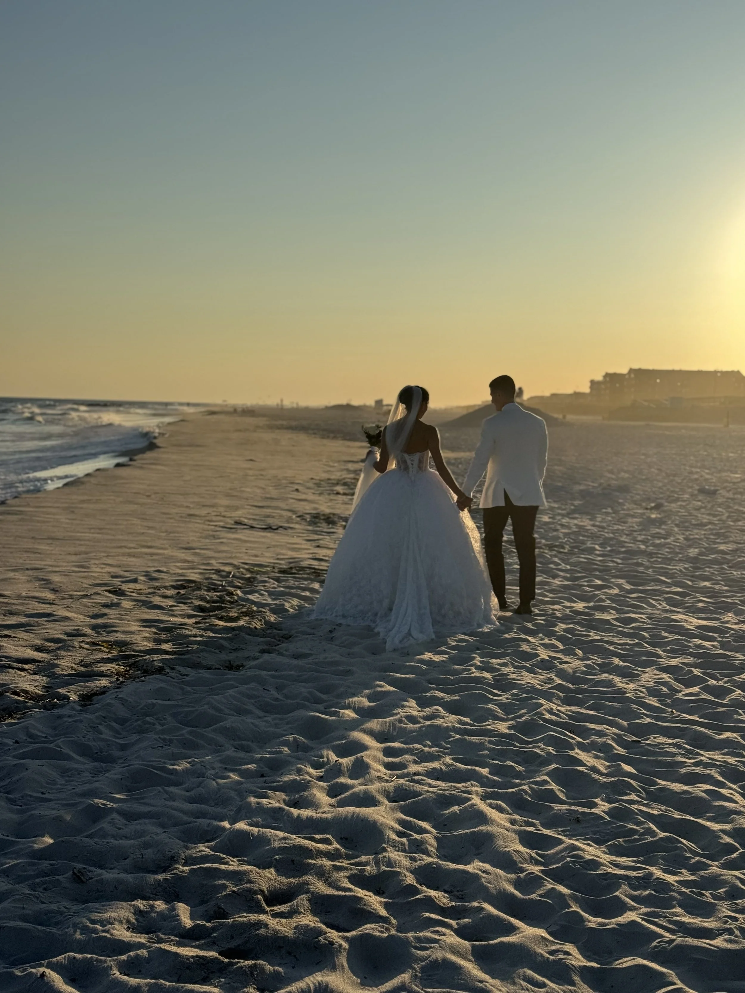 A bride and groom holding hands walking on a beach during sunset.