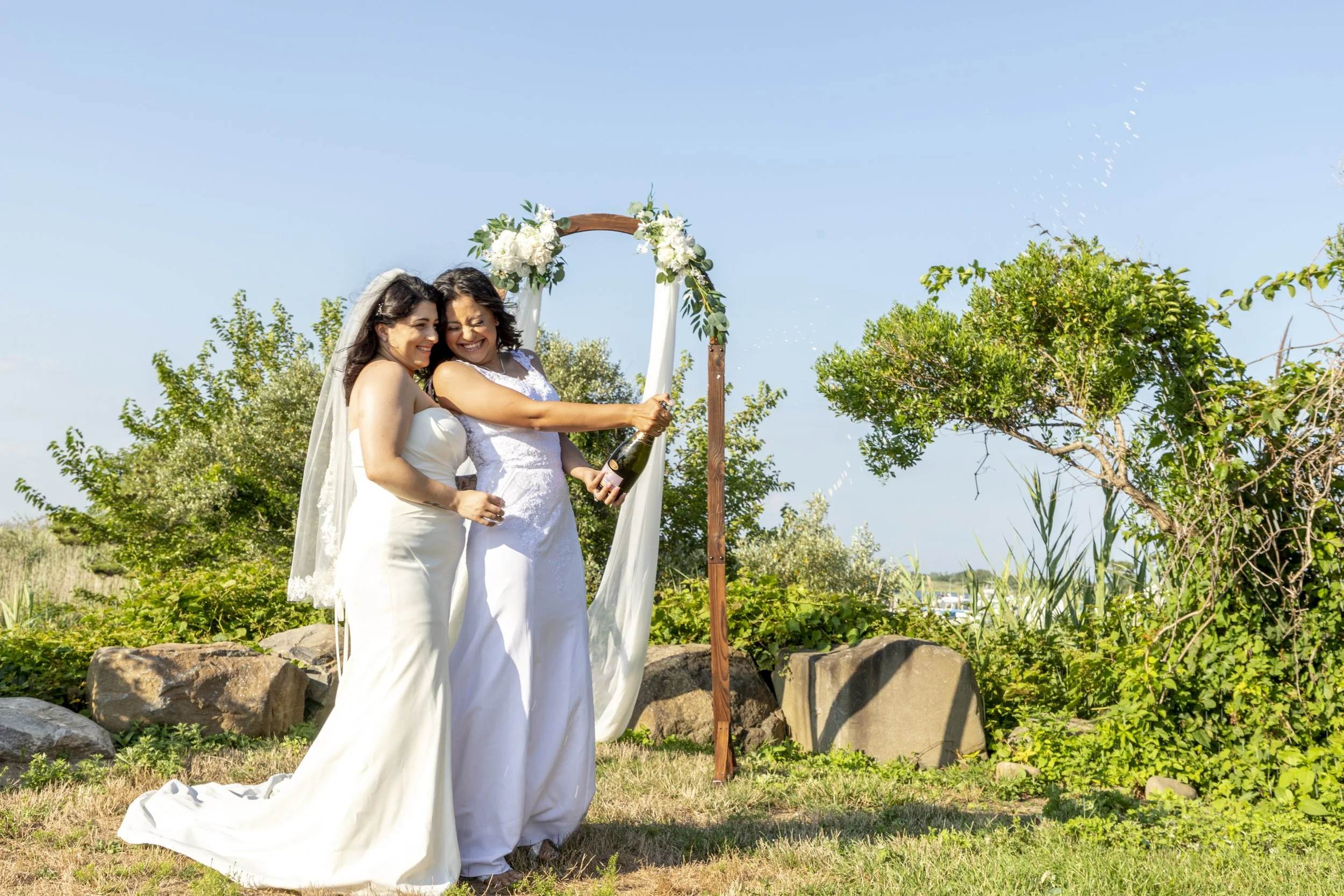 Two women in wedding dresses celebrating outdoors during daytime, one of them opening a champagne bottle with a splash, near a decorative arch adorned with flowers and greenery, with trees and rocks in the background.