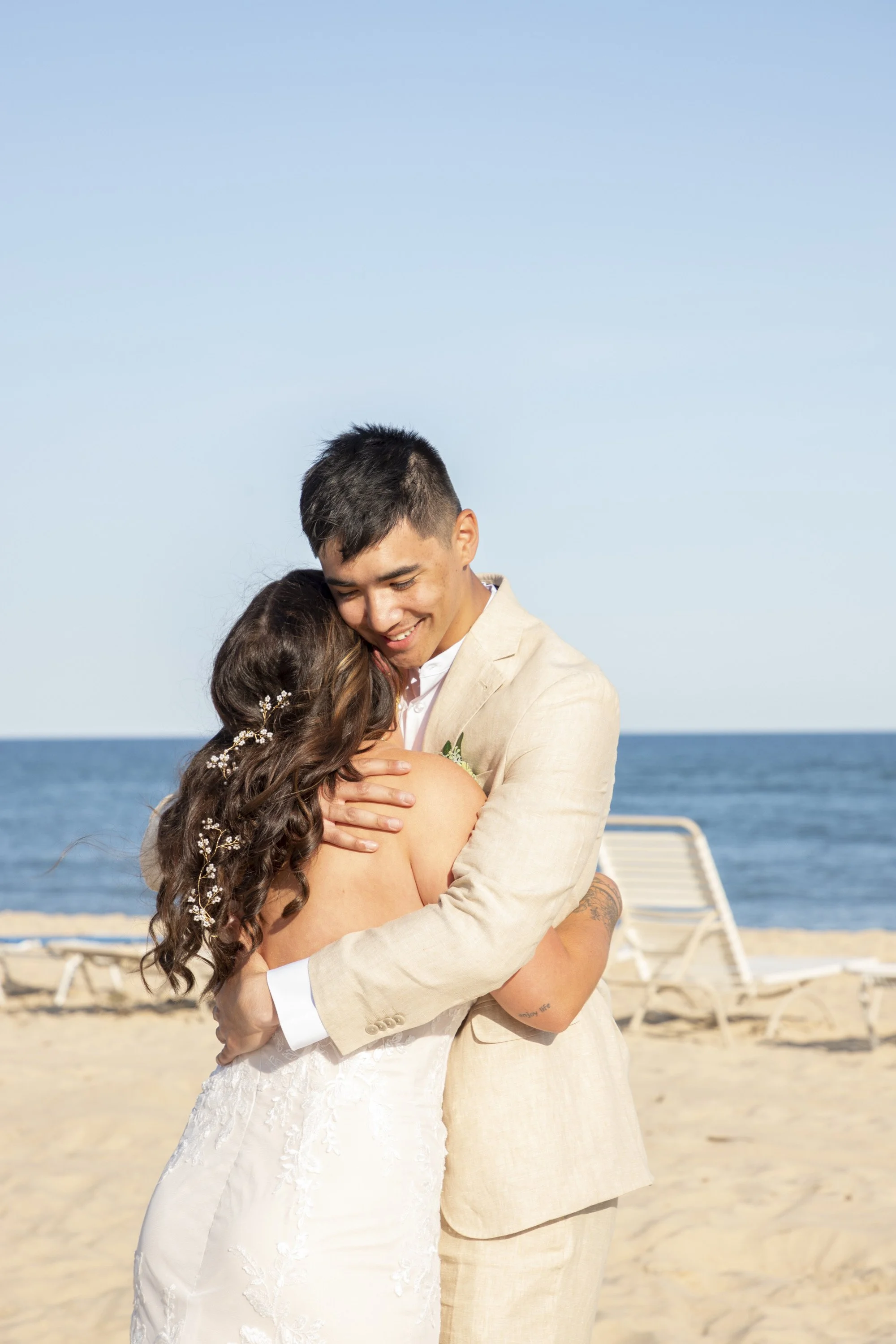 A couple hugging on the beach, the man dressed in a beige suit, the woman in a white wedding dress, with Lounge chairs and the ocean in the background.