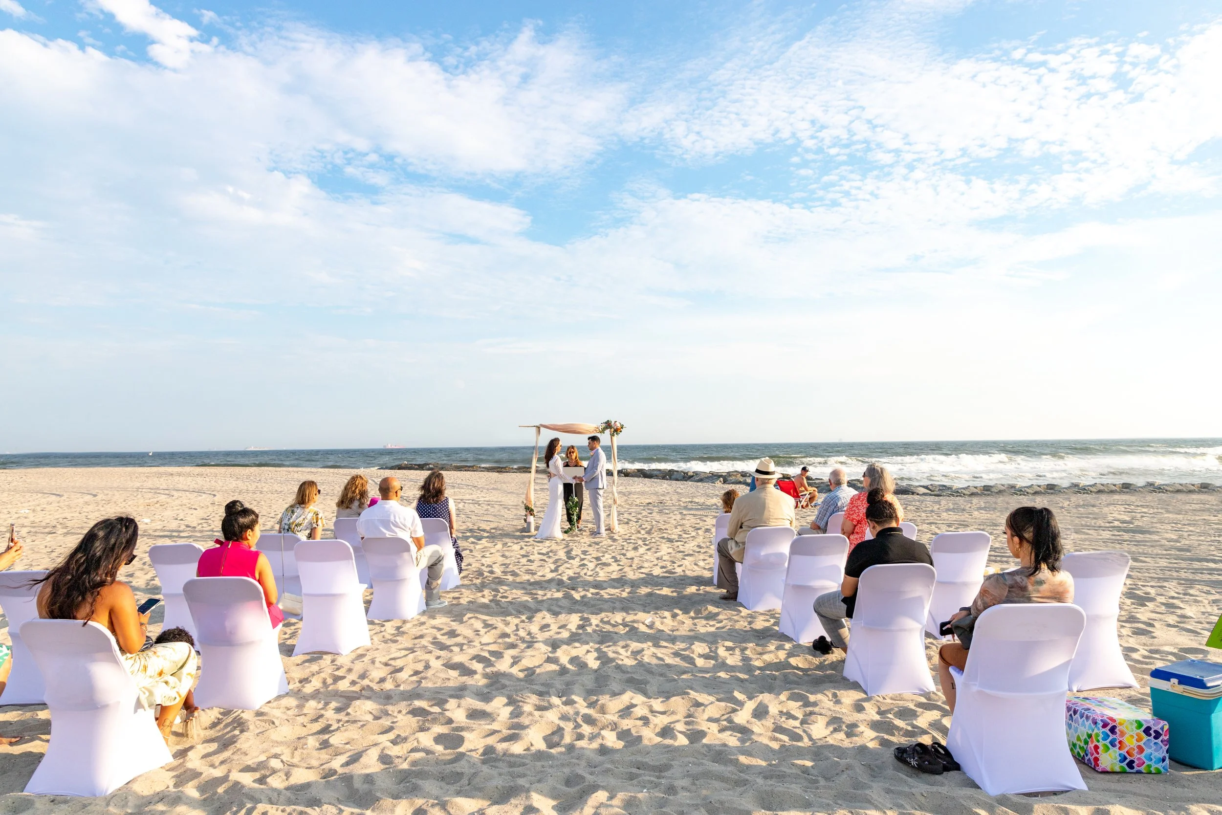 A beach wedding ceremony with an arch and chairs arranged on the sand, facing the ocean, under a partly cloudy sky.
