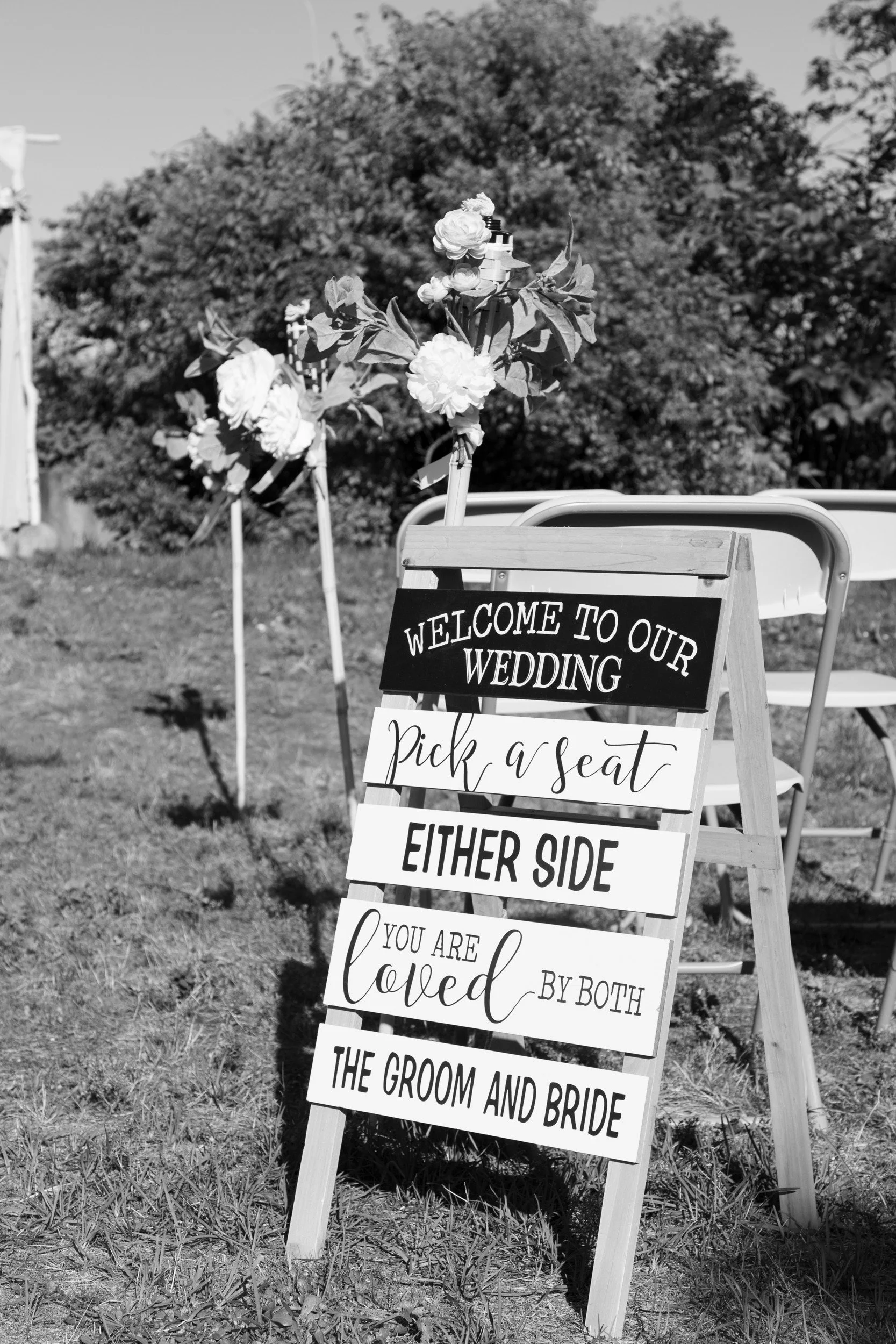 Wooden ceremony welcome sign on the lawn reading 'Pick a seat, either side.'