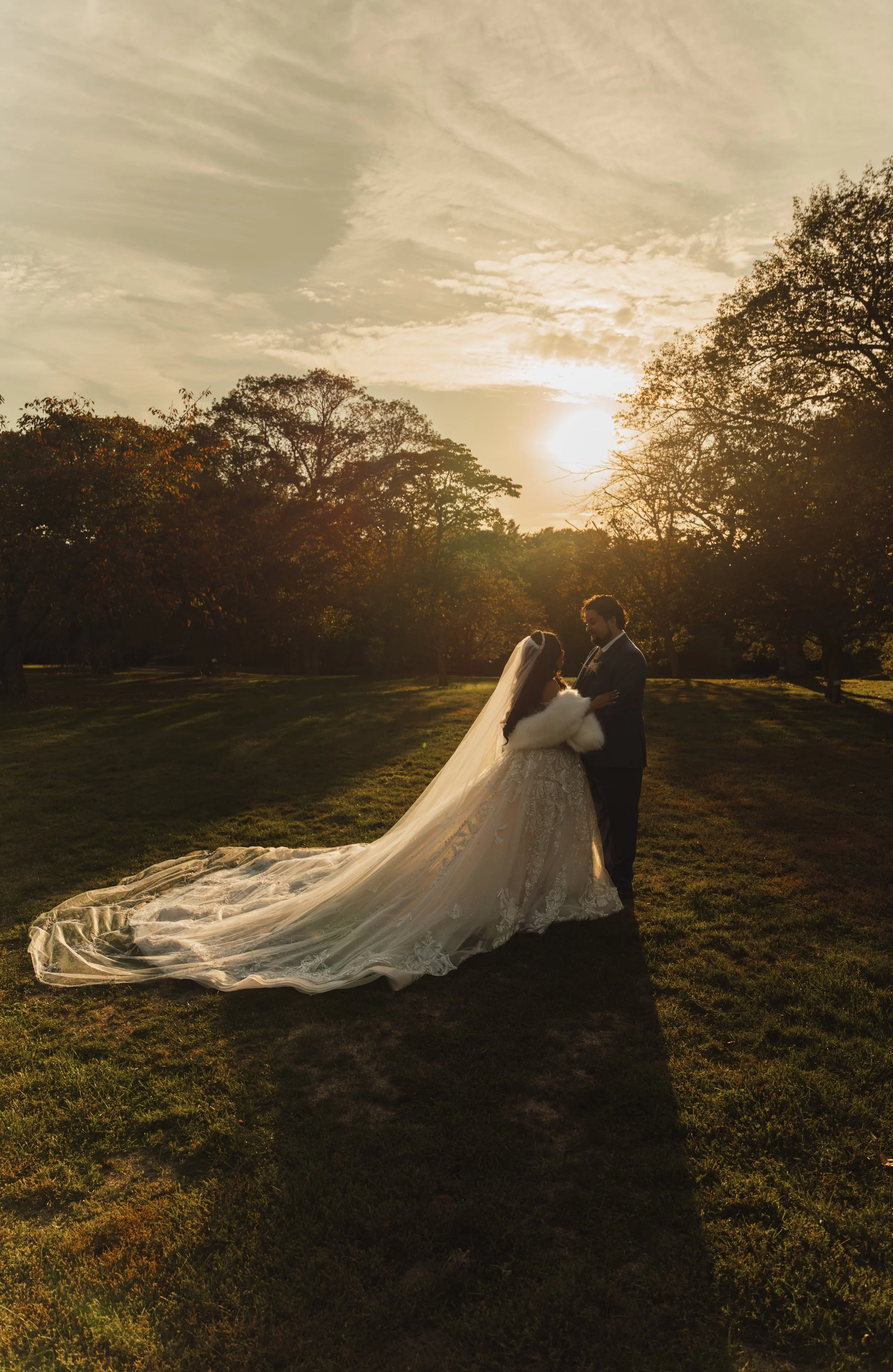 A bride and groom standing close together in a park during sunset, with trees and a cloudy sky in the background. The bride is wearing a white wedding dress with a long train and veil, and the groom is in a dark suit. The sunlight creates a warm glow around them.