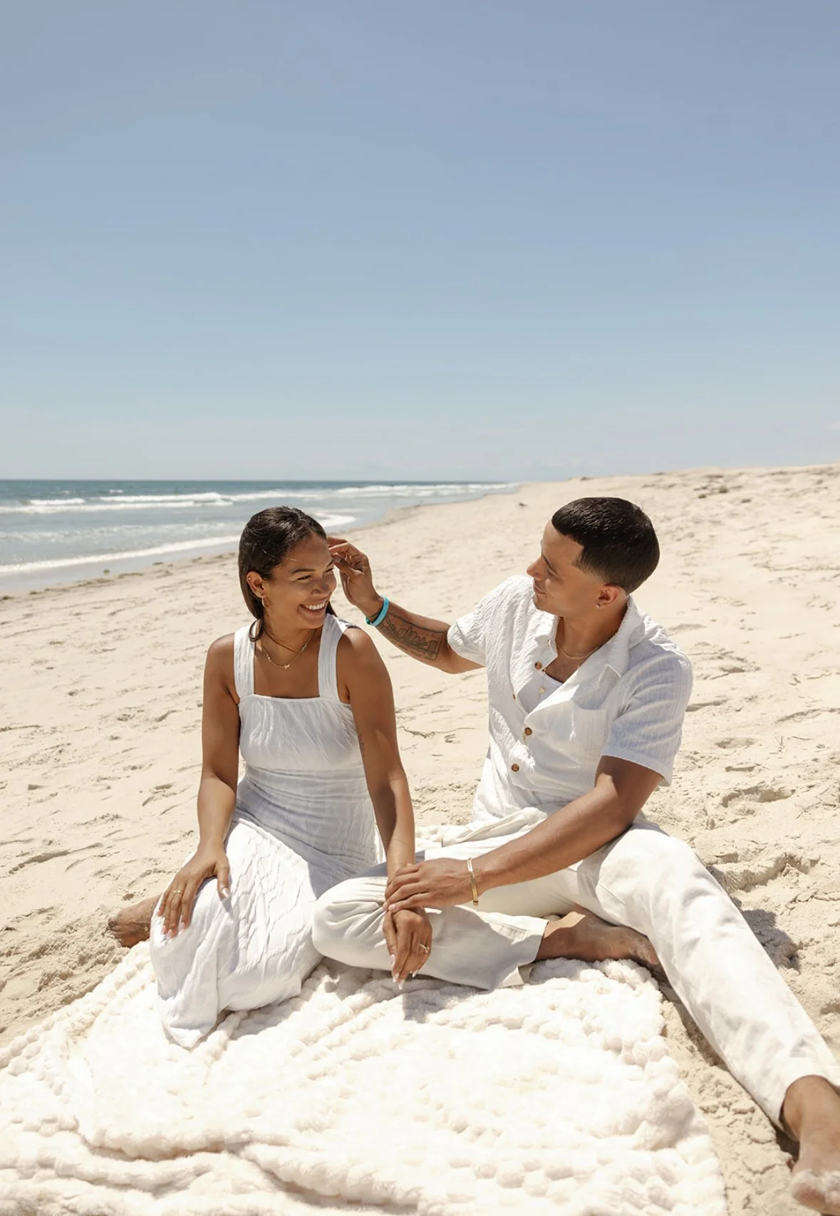 A couple sitting on a blanket on the beach, smiling and interacting with each other, with the ocean and sky in the background.