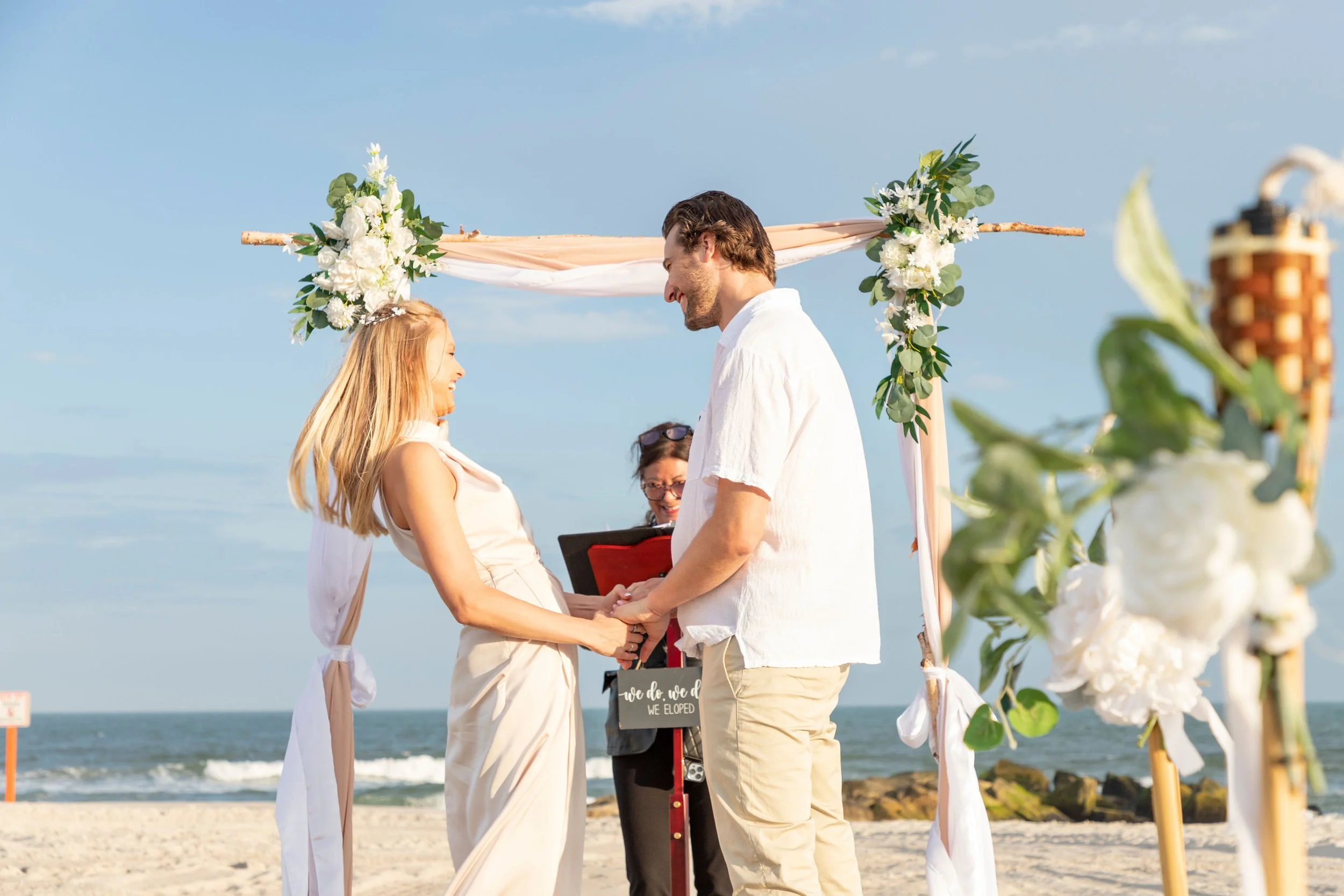 A couple getting married on the beach with an officiant holding a book, under a wooden arch decorated with white flowers and green foliage, with the ocean and sky in the background.