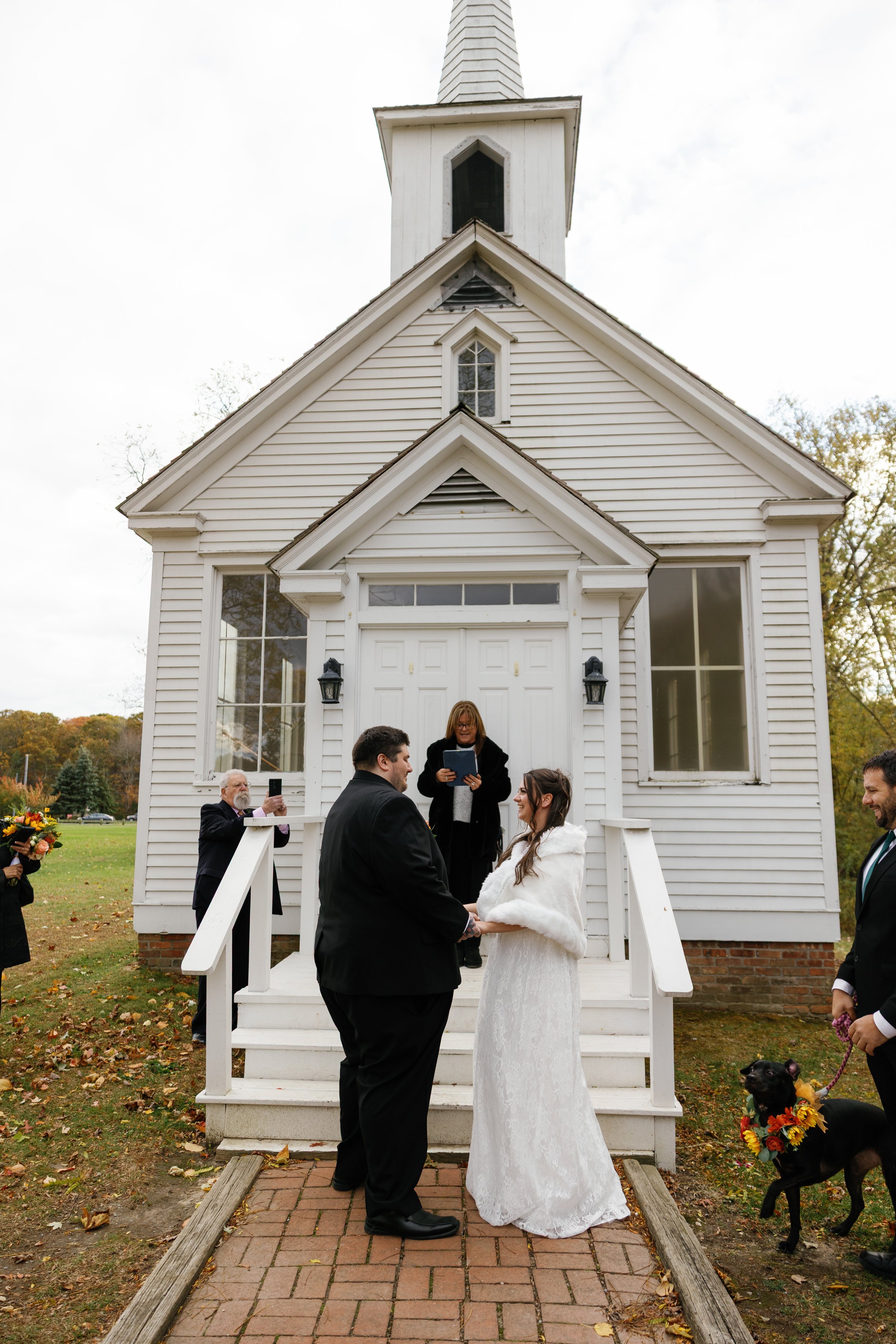 A wedding ceremony taking place outside a white church with a steeple. The bride and groom stand on the brick path, holding hands, with the officiant behind them. Guests are present, one taking photos and carrying flowers, and a dog with a colorful floral collar is also part of the ceremony.