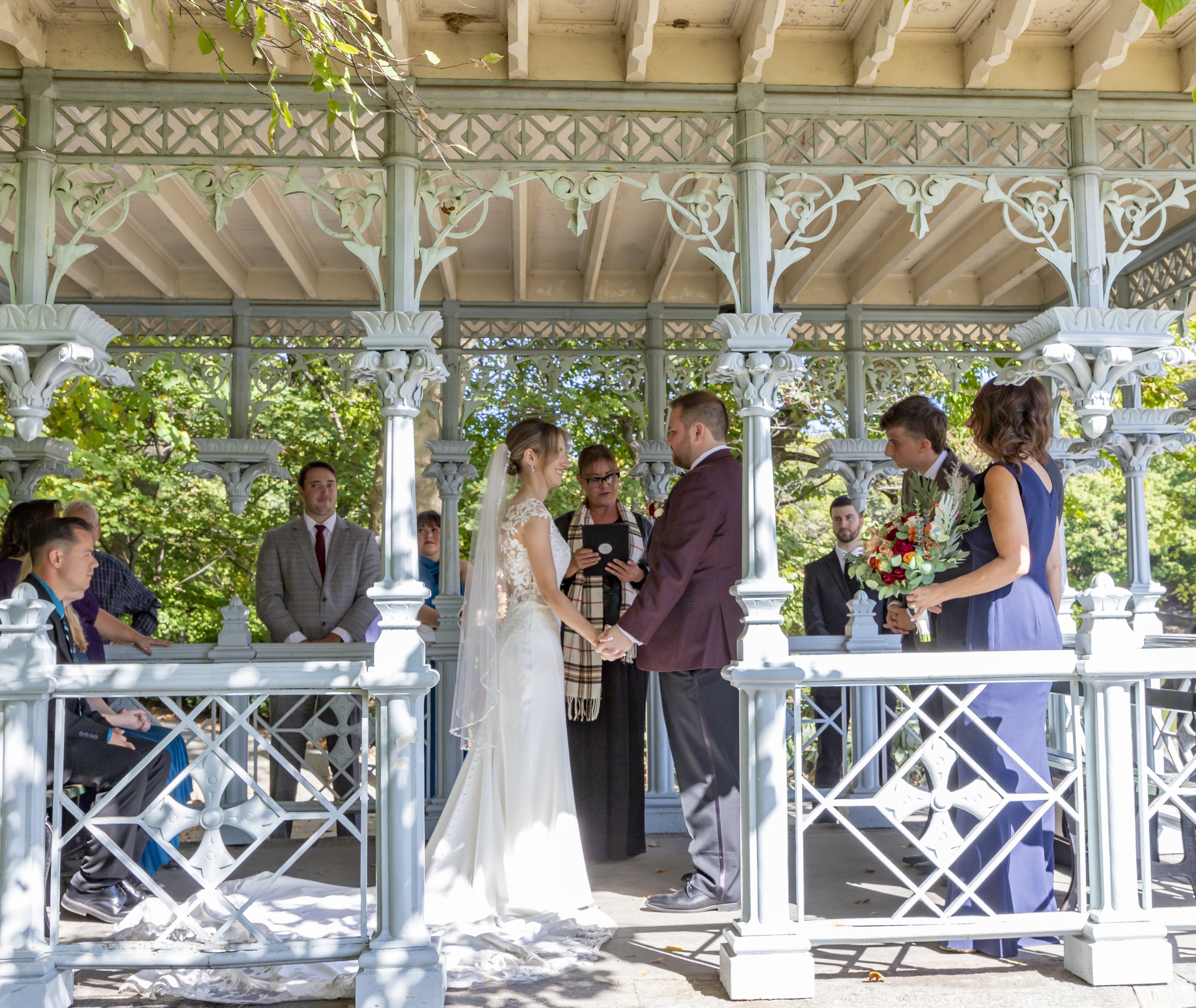 A wedding ceremony taking place in a decorative, outdoor gazebo surrounded by greenery. The bride and groom are holding hands and facing each other as they exchange vows, with an officiant between them. The bride is in a white wedding dress with lace details, and the groom is in a maroon jacket and gray pants. Bridesmaids and groomsmen, some holding bouquets, are standing and sitting nearby.