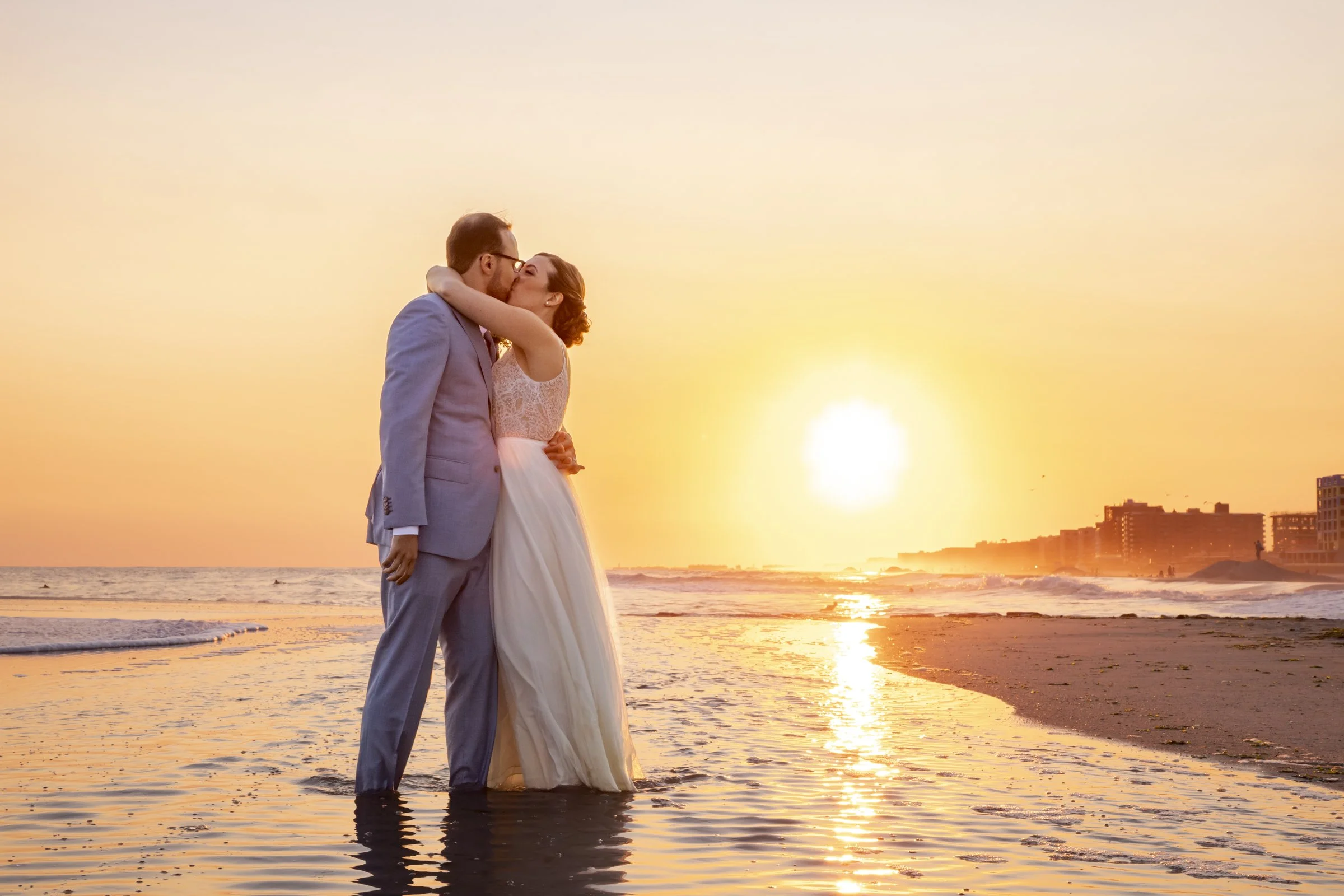 A couple in wedding attire sharing a kiss at sunset on the beach, with the ocean and city buildings in the background.