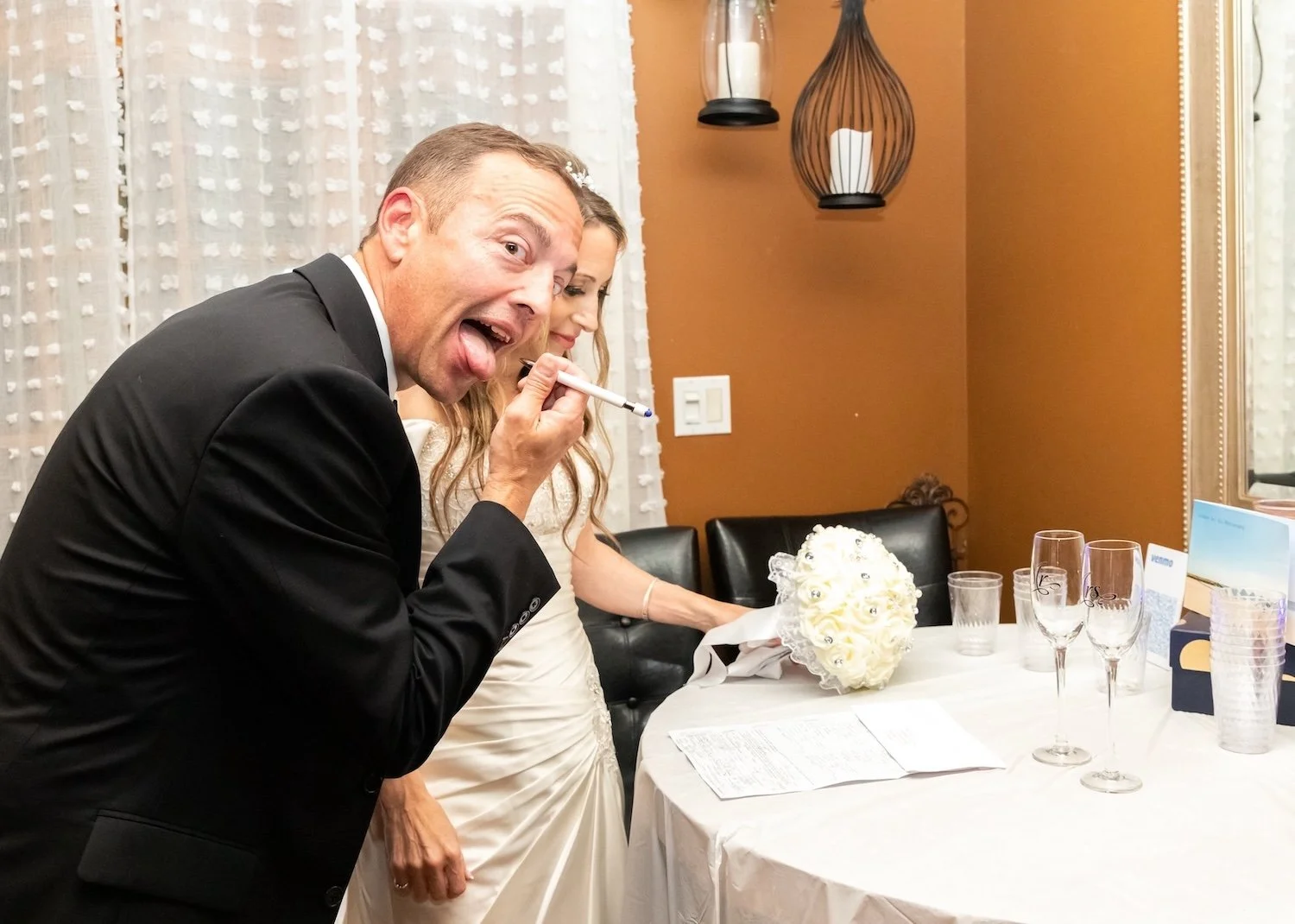 Man in a tuxedo making a silly face with his tongue out, holding a pen near his mouth at a wedding reception table, with a bride in a white dress sitting beside him, table decorated with a white bouquet, glasses, and a guest book in the foreground.